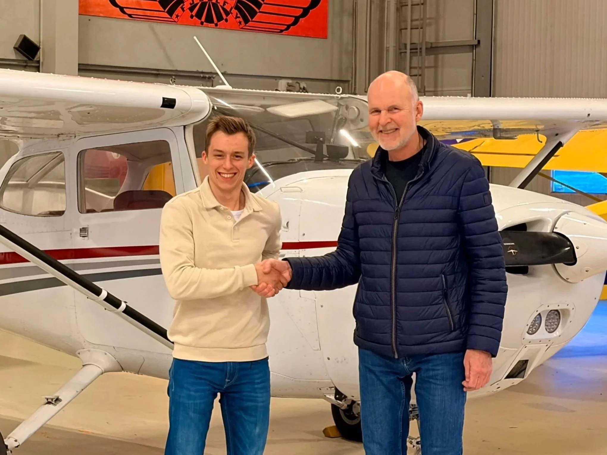 To menn står og smiler mens de håndhilser foran en liten fly i en hangar.