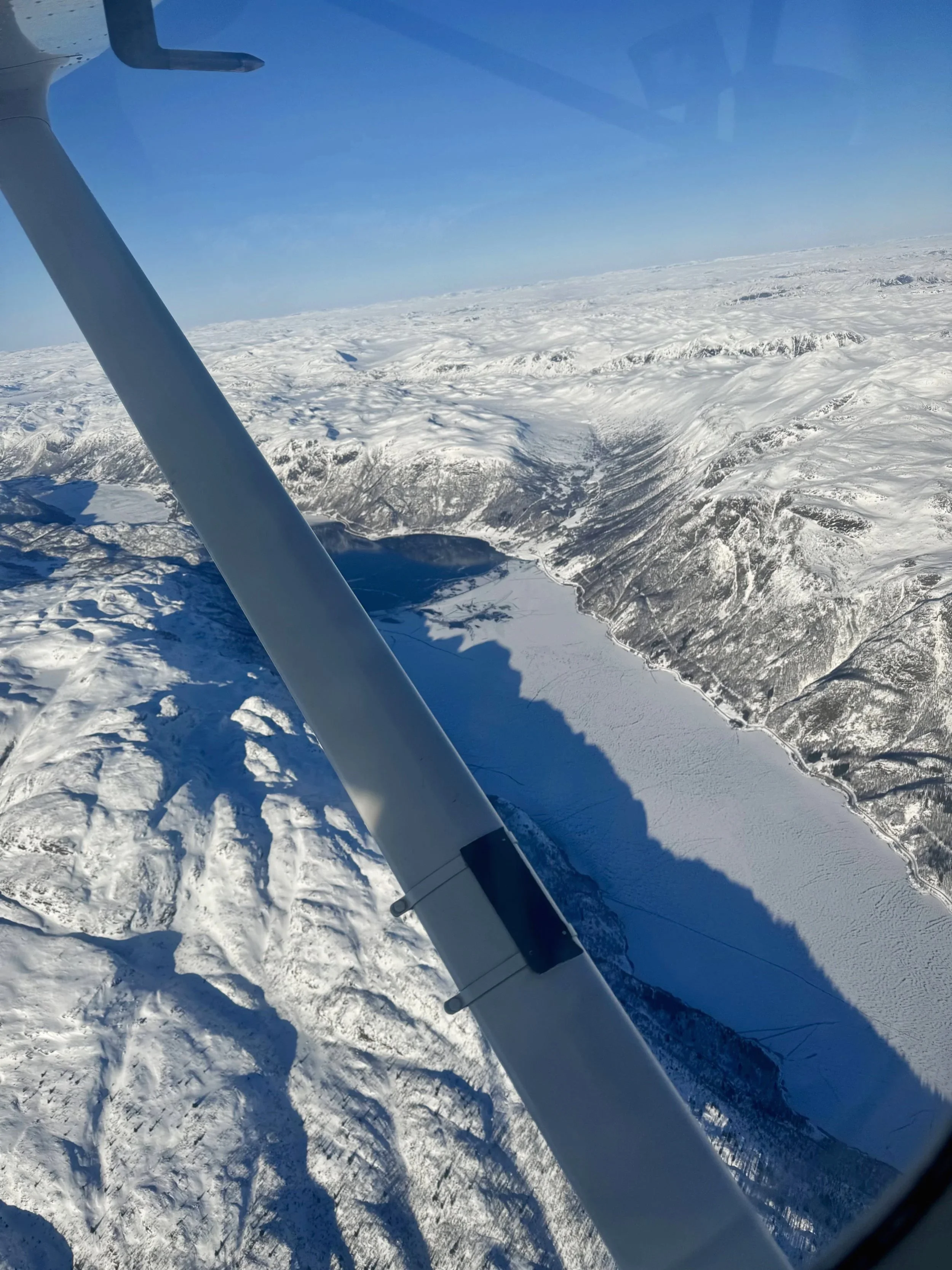 Flyfoto av snødekte fjell, innsjø og kalde landskap sett fra en liten flykabin, med en flyvinge i forgrunnen.