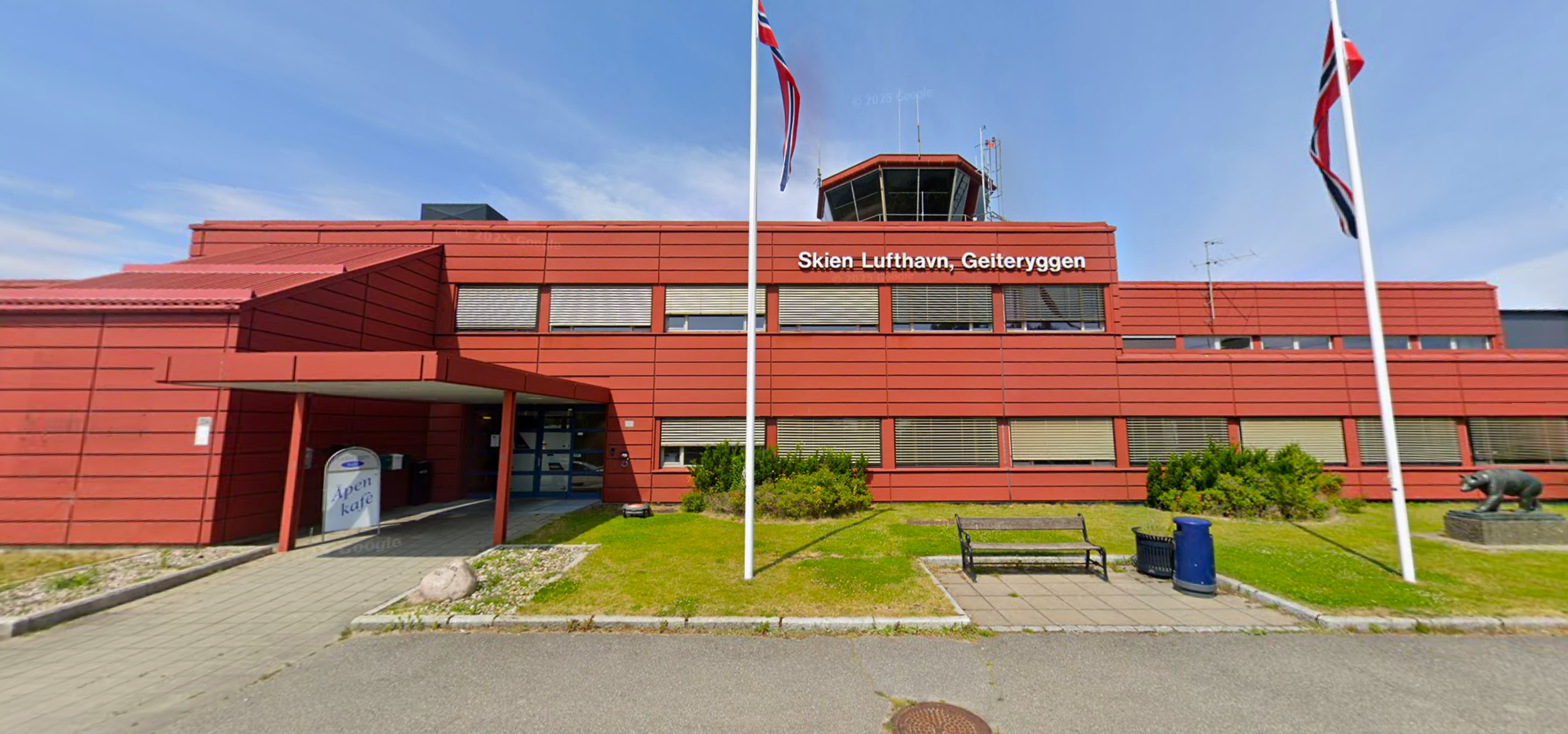 Utsiden av bygningen Skien Lufthavn, Geiteryggen, med en rød bygning, flaggstenger med norske flagg, en benk, søppelkurv, og en skulptur av en bjørn foran bygningen.