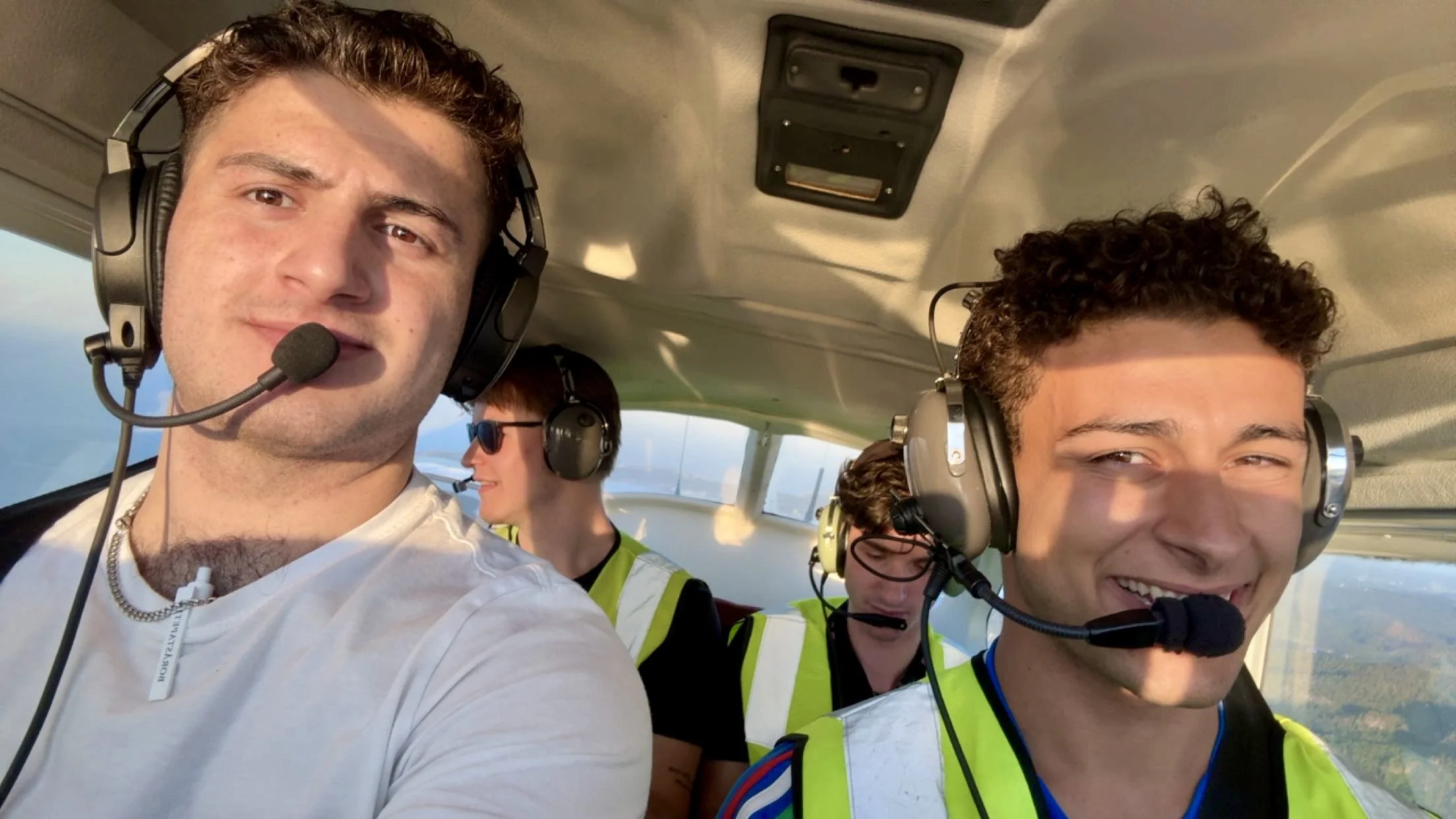 Four young people flying in an airplane cockpit, wearing headsets with microphones, with two men in the foreground and two women in the background, smiling and enjoying the flight.