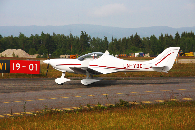 A small white propeller aircraft with red stripes on a runway, with a forested background and mountain range in the distance.