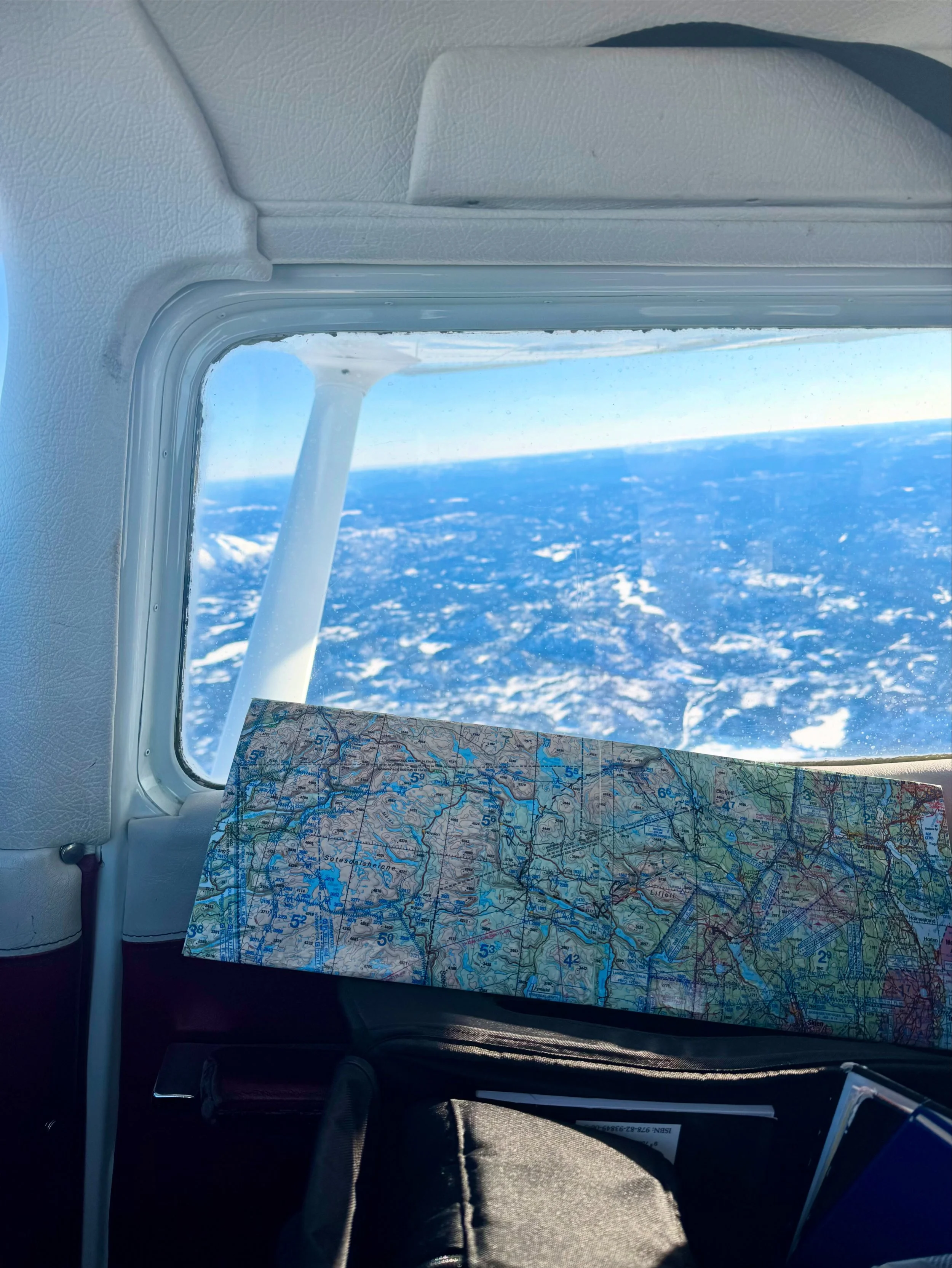 View from inside an aircraft showing a window with snowy land and ocean below. A map is placed on a seat.