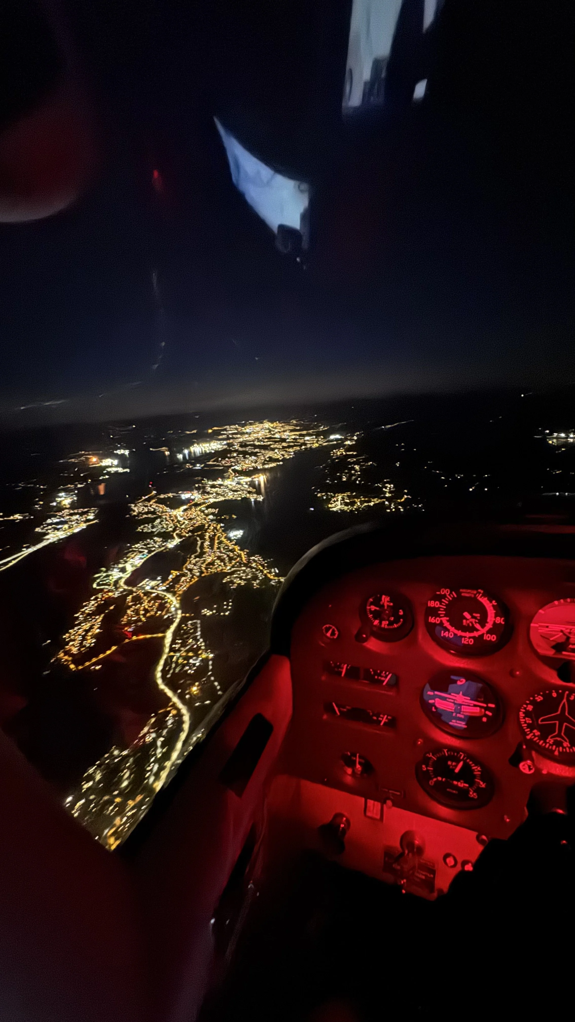 Nighttime aerial view from inside an aircraft cockpit showing illuminated city lights below and the dashboard instruments lit in red.