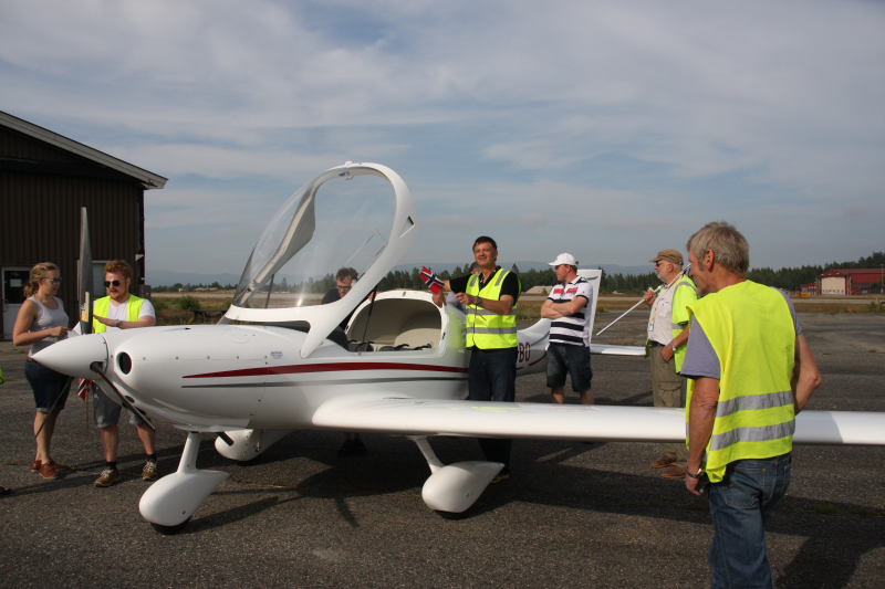 Group of people on an airfield gathered around a small white aircraft with a transparent canopy, with one person holding a model airplane, under a partly cloudy sky.