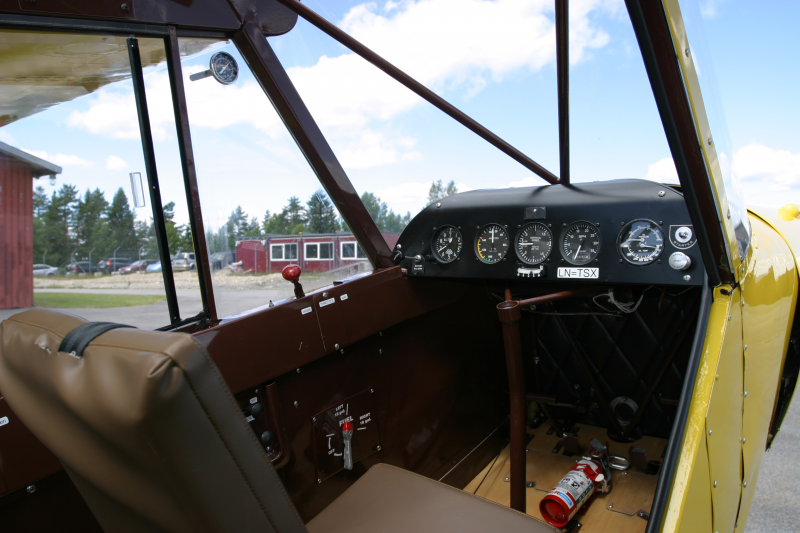 Interior of a small yellow vintage aircraft cockpit with several gauges, a control stick, and a fire extinguisher on the floor.
