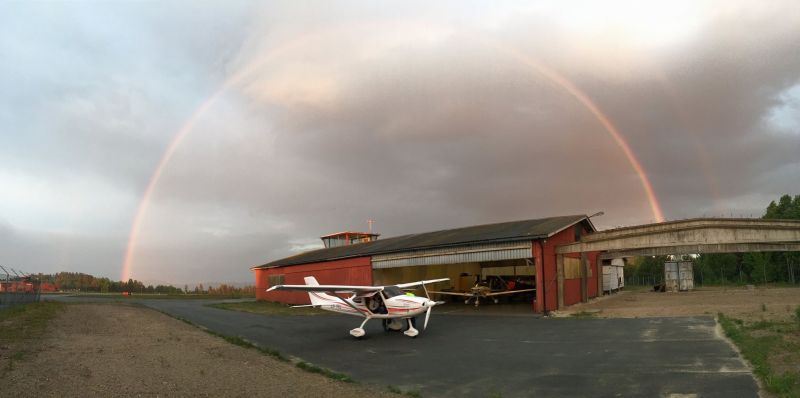 Small airplane parked outside a hangar at an airfield with a double rainbow arched in the cloudy sky.