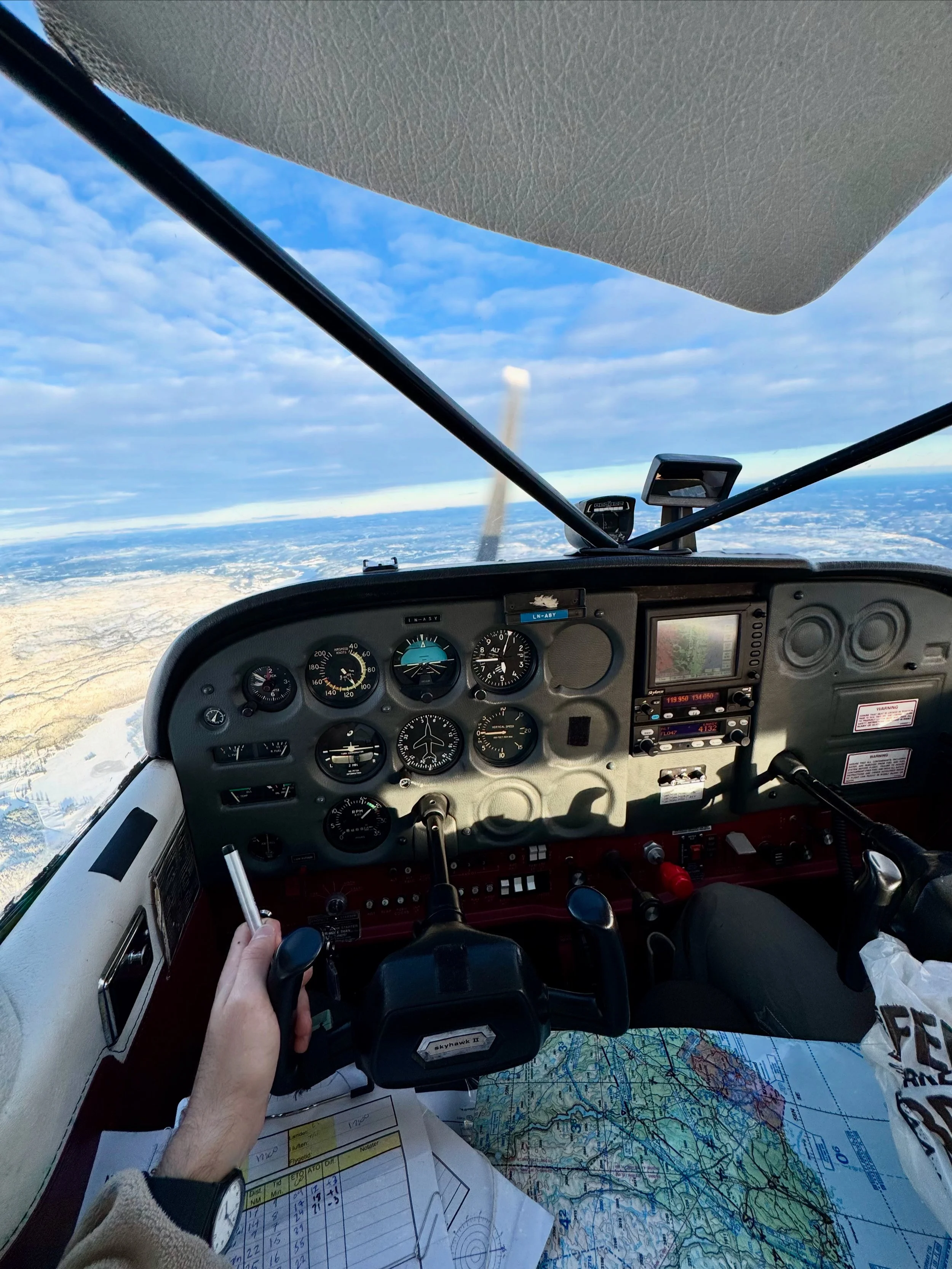 Interior view of a small aircraft cockpit during flight, showing the main instrument panel with gauges, a hand holding a control yoke, and a view of the sky and landscape below through the windshield.