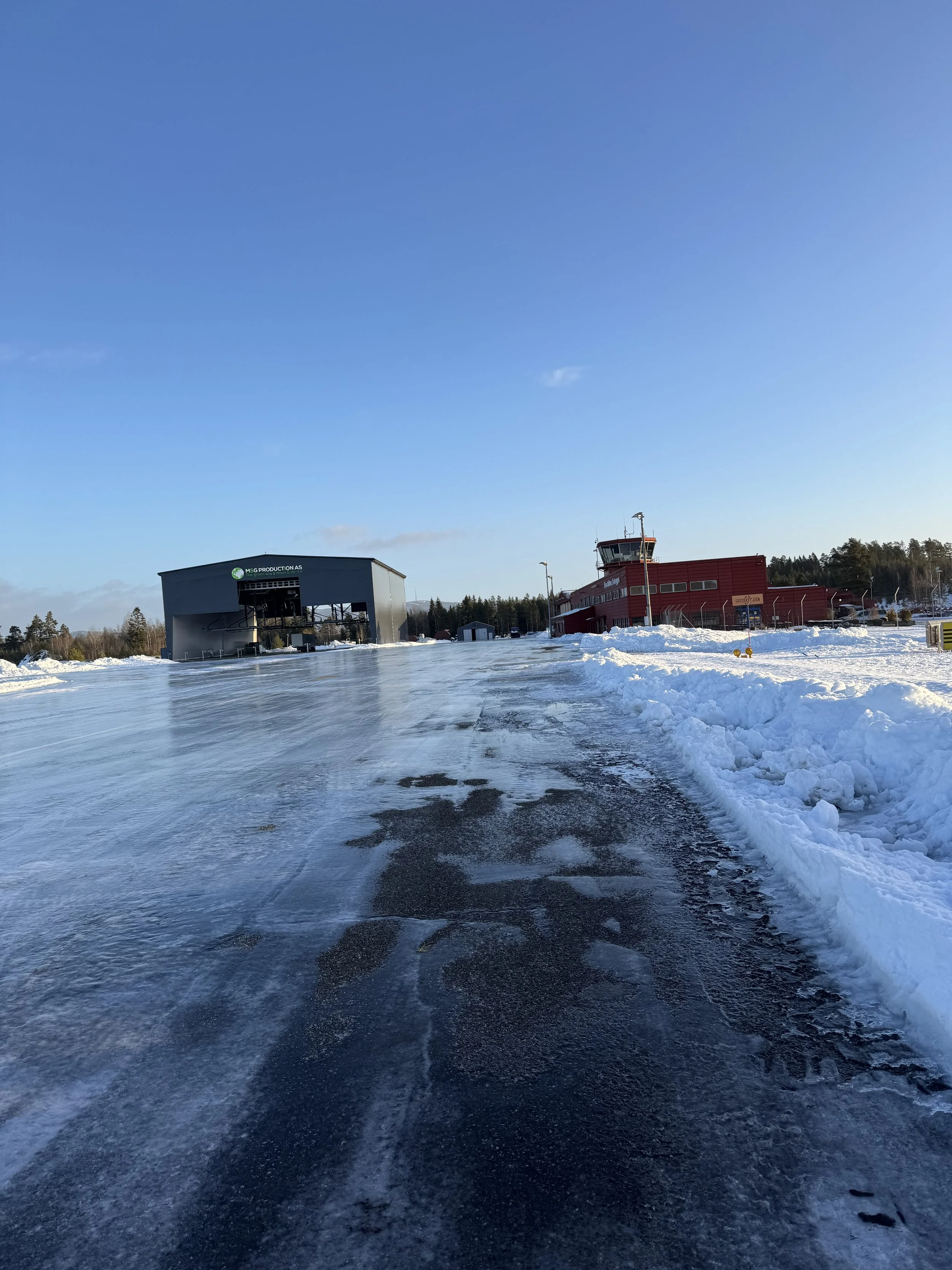 Vinterlandskap med en islagt flystripe, to hangarer og flykontrolltårn. Klar blå himmel.