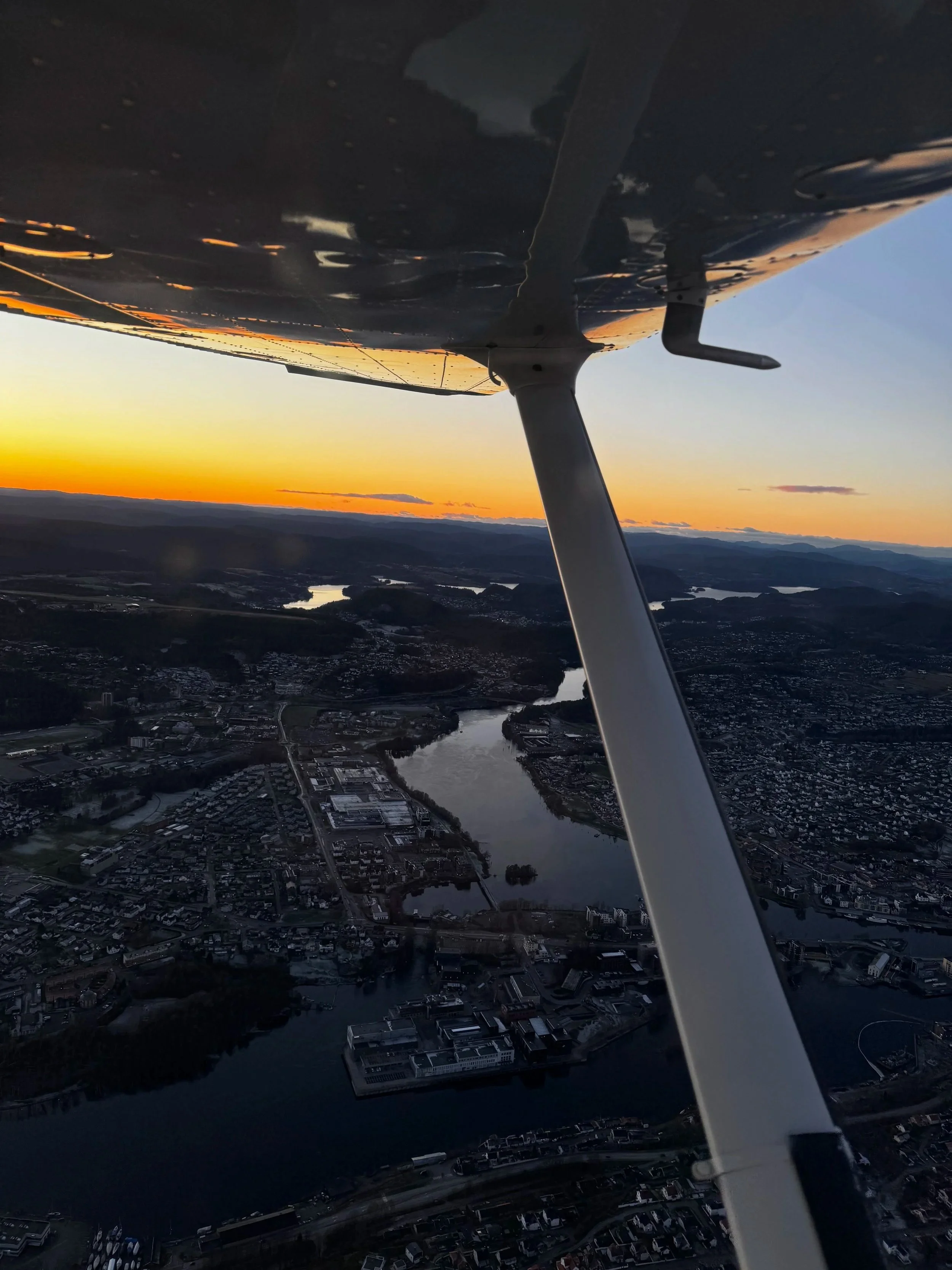 Flyfoto av en by med vann, tatt fra et fly under solnedgang, med del av flyets vinge og stangen i forgrunnen.