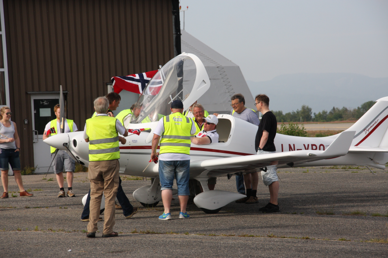 Group of people gathered around a small white airplane with red trim, some in yellow safety vests, with a British flag displayed, on an airfield with hangar and mountains in the background.