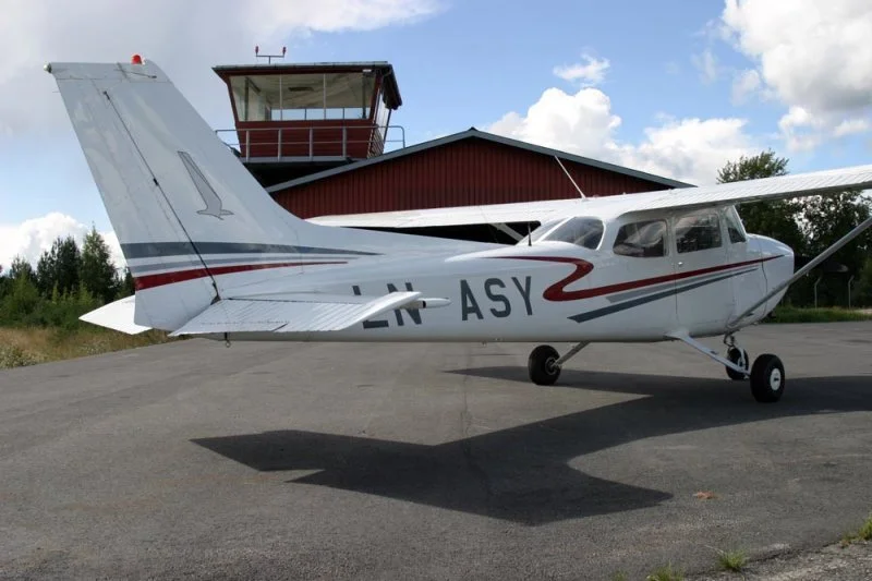 Small white aircraft with red and gray stripes parked on tarmac near red airport control tower and hangar, with blue sky and clouds in the background.
