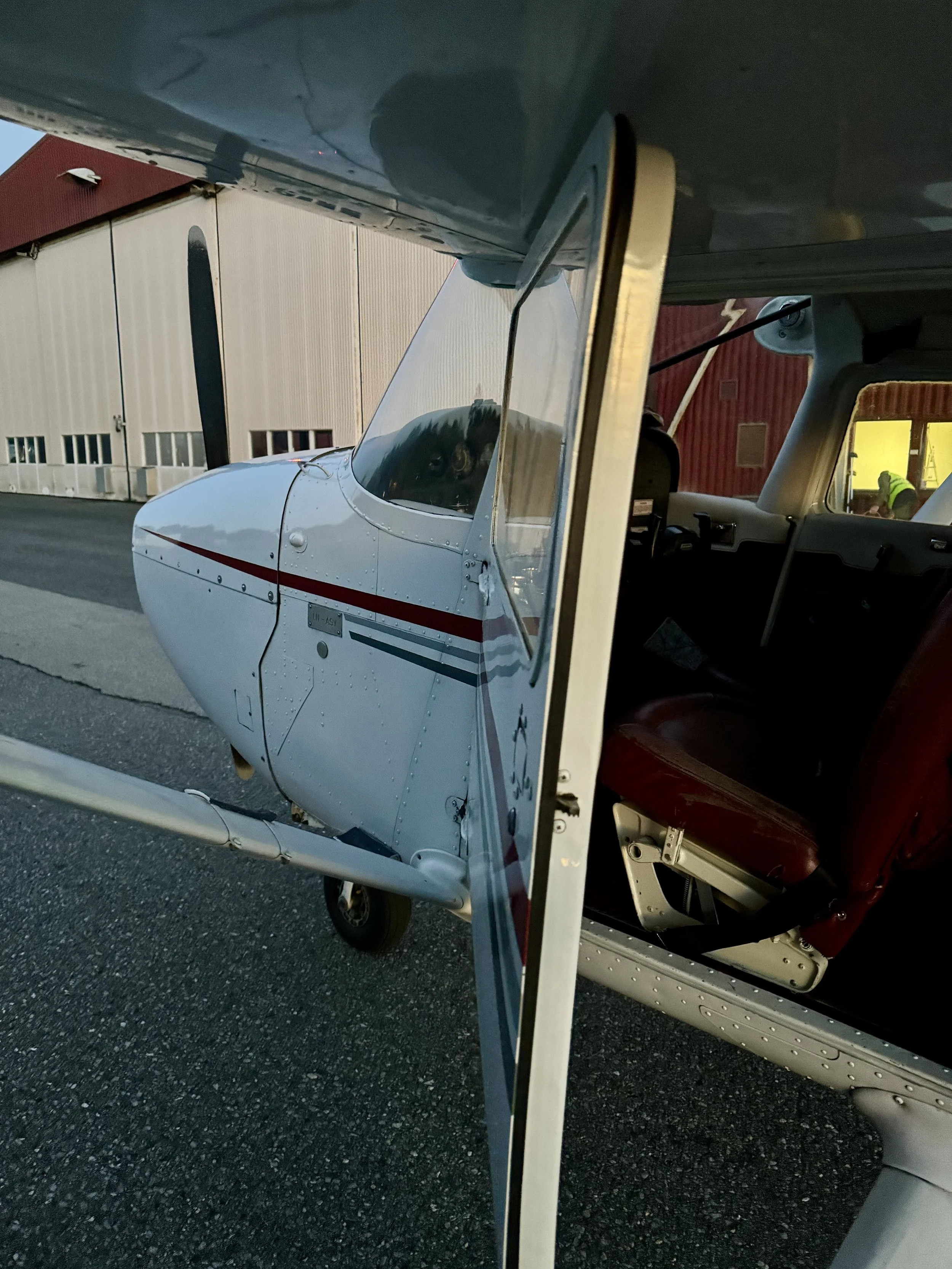 Smal sportskrogfly med cockpit og ensengsseter, parkert på asvalt med hangar i bakgrunn.