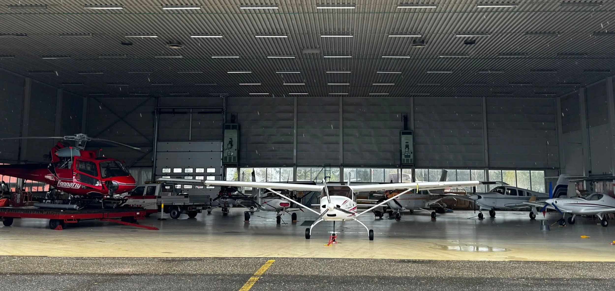 Innendørs bakke hangar med flere små fly og helikopter parkert, store vinduer og en høy tak.