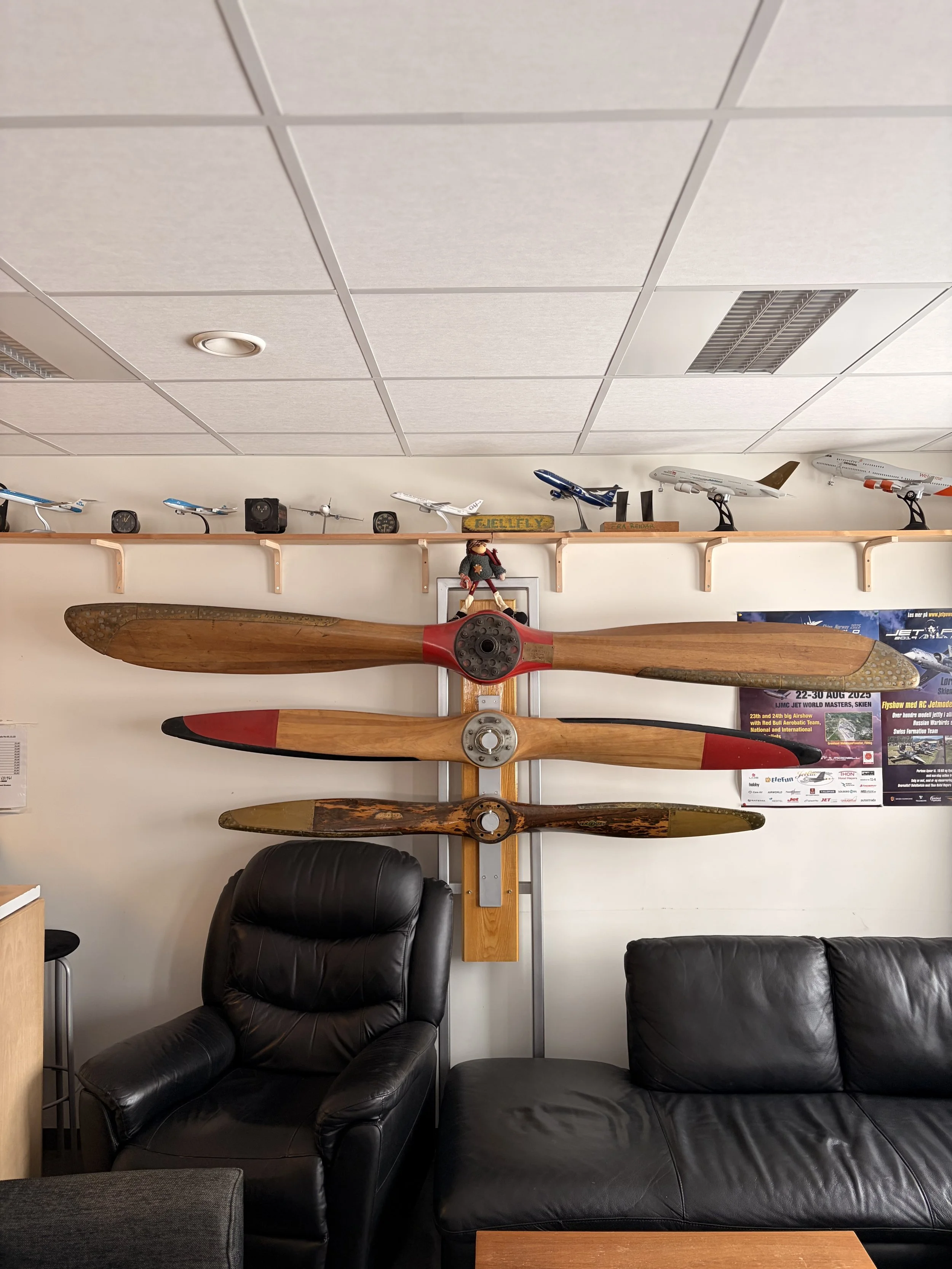 Wall display with three vintage airplane propellers mounted vertically, a model airplane collection on a shelf above, and a seating area with black leather chairs.
