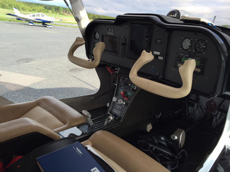 The cockpit of a small airplane with two control sticks, digital displays, and gauges. Outside the window, another small aircraft is parked on the tarmac with a runway and trees in the background.