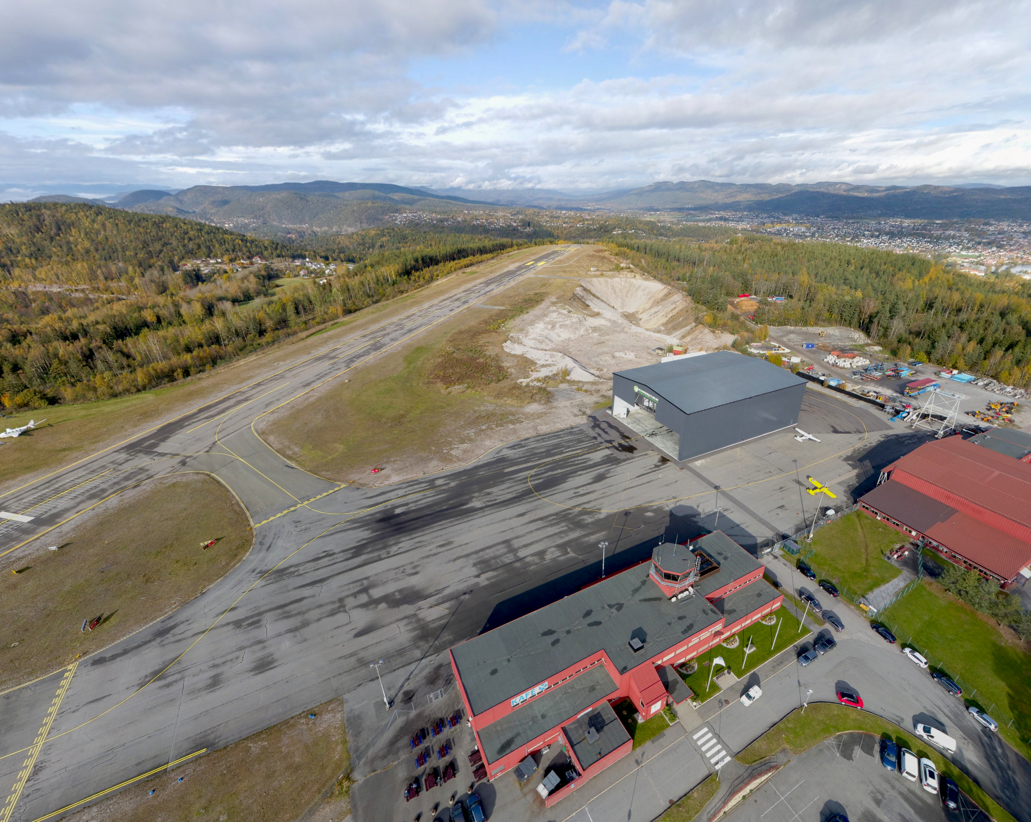 An aerial view of a small airport with runways, surrounding forest, and a red building. There are some aircraft and equipment near the building, with a parking lot in front.