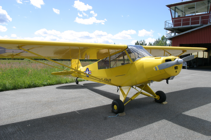 Yellow vintage plane with black wheels parked on tarmac near a red building with glass windows and a balcony.