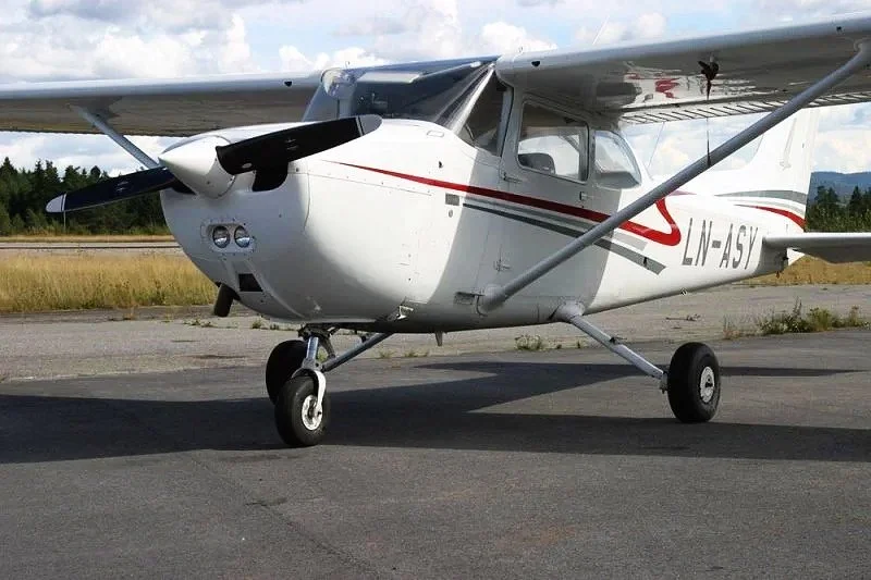 Small white single-engine propeller airplane parked on the tarmac with mountains and grass in the background.