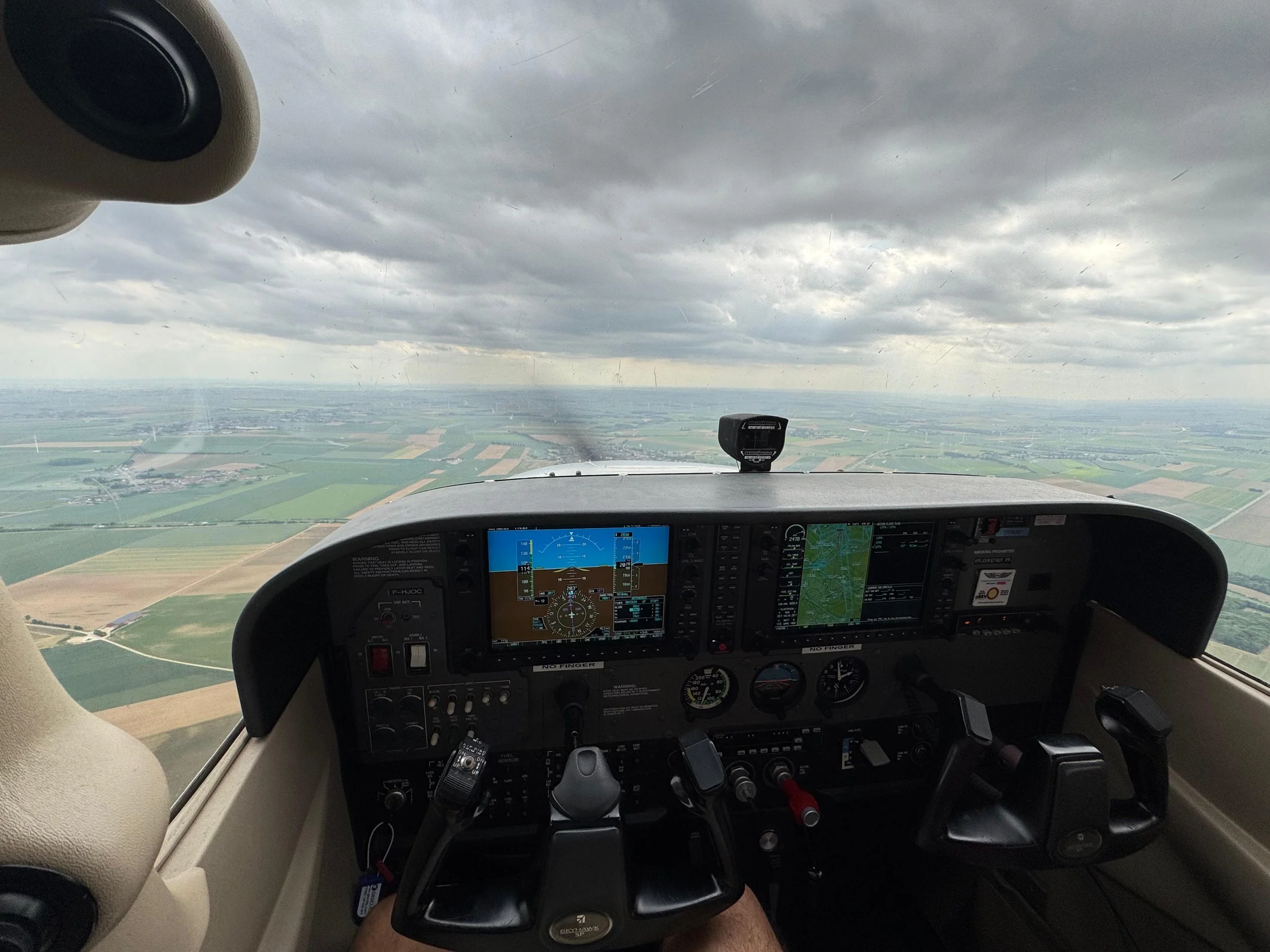 Inside view of a small airplane cockpit with flight instruments and navigation screens, flying over farmland on a cloudy day.