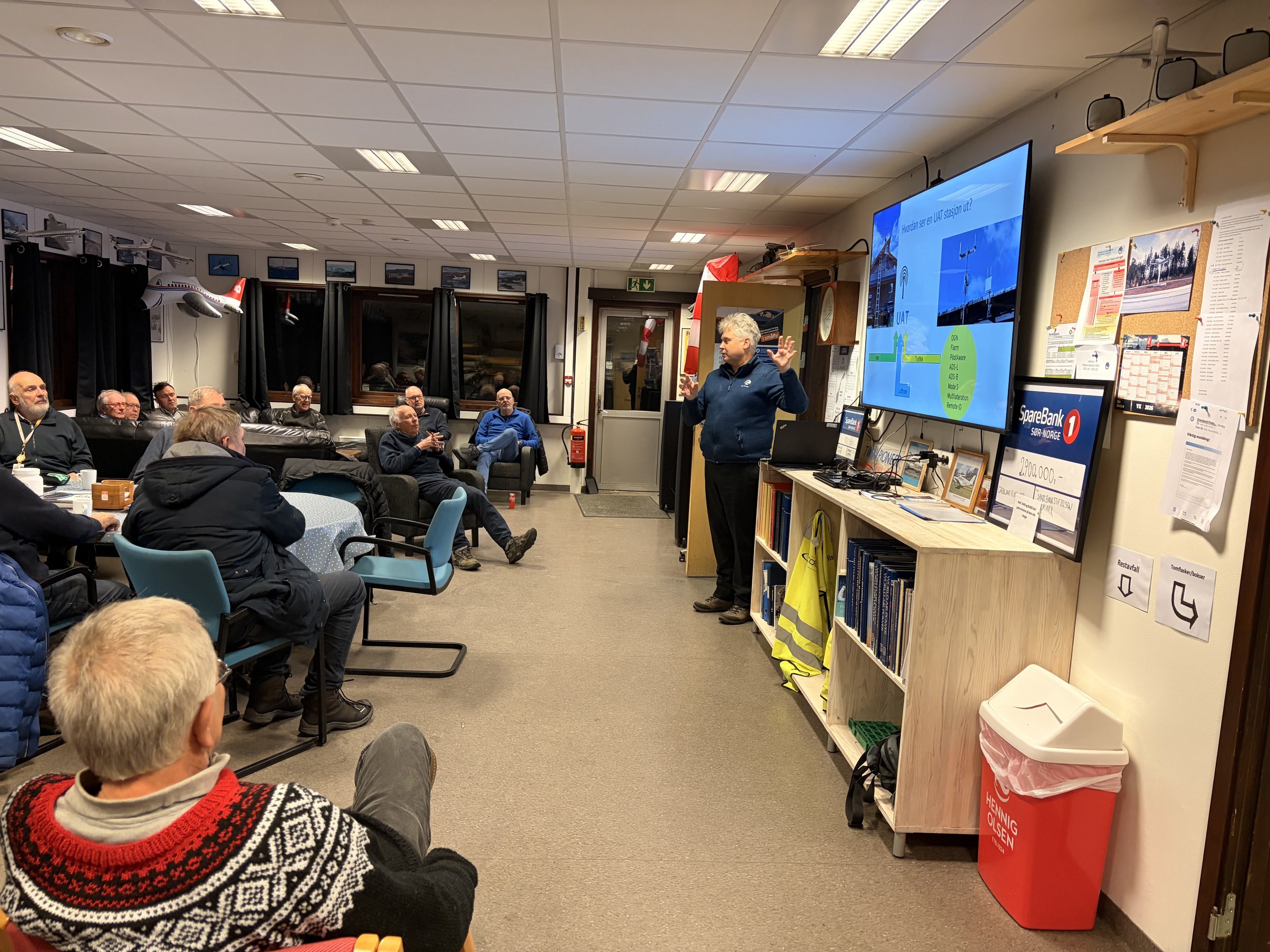 A group of people sitting in a room watching a man giving a presentation. The room has a TV screen displaying a slide with a map and text. The audience appears attentive, and some are taking notes. The setting looks like a community meeting or seminar.