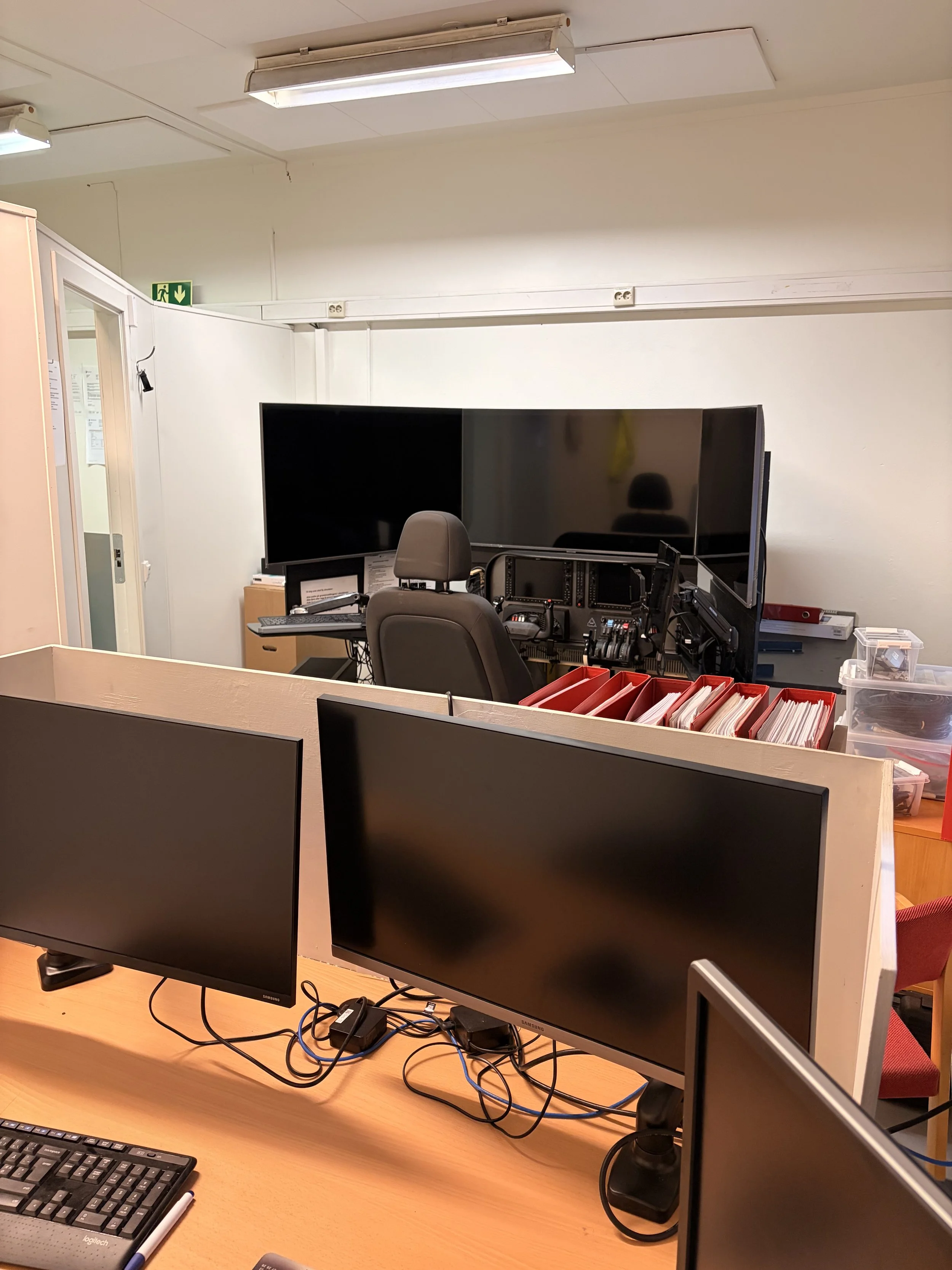 Office workspace with multiple computer monitors, a control desk with screens, and filing trays, viewed from behind a partially obscuring divider.