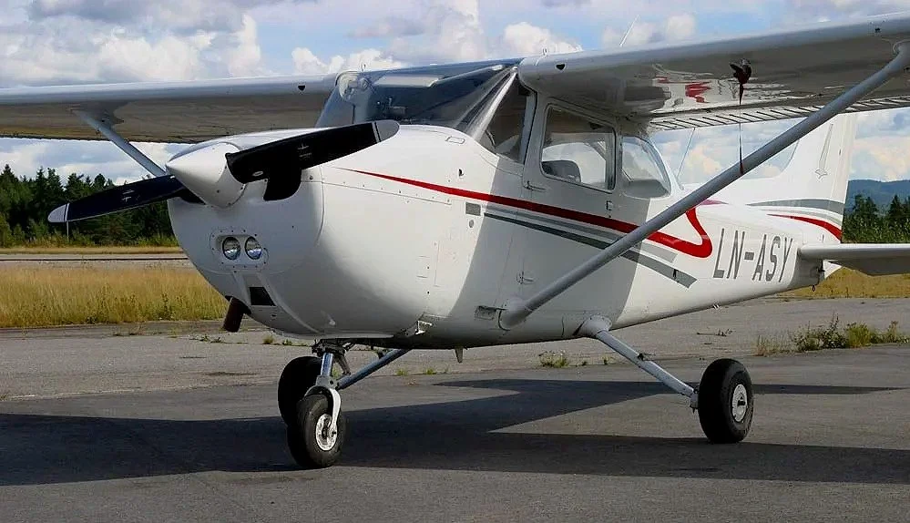 Small white single-engine propeller airplane parked on runway with grassy field and trees in background.