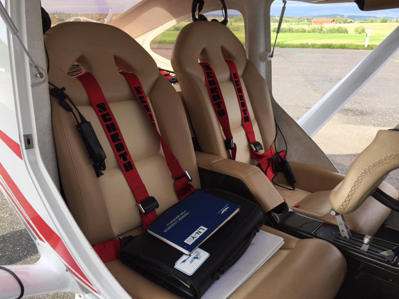 Interior of a small airplane with two beige leather seats equipped with red Sparco harnesses, an aircraft manual, and a black case on one seat.