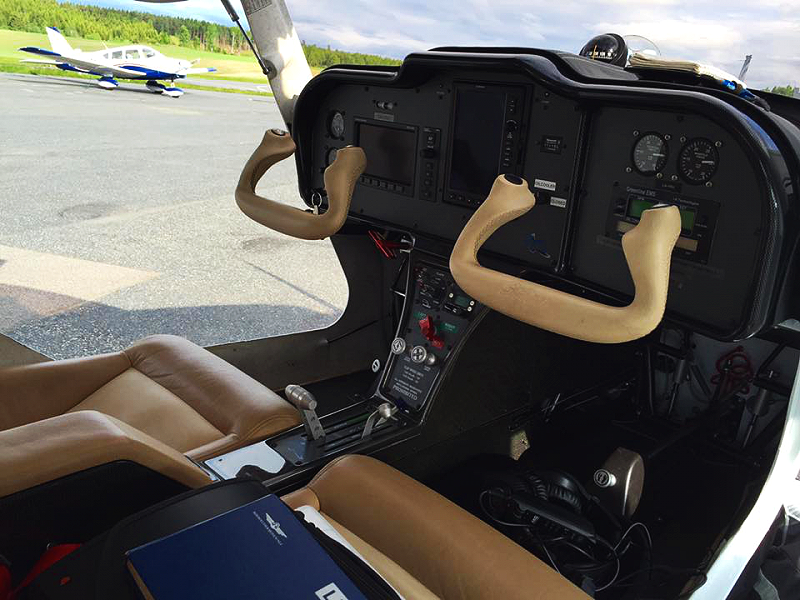 Interior view of a small aircraft cockpit with two brown seats, beige control sticks, and various instruments, with a small airplane visible on the tarmac outside.