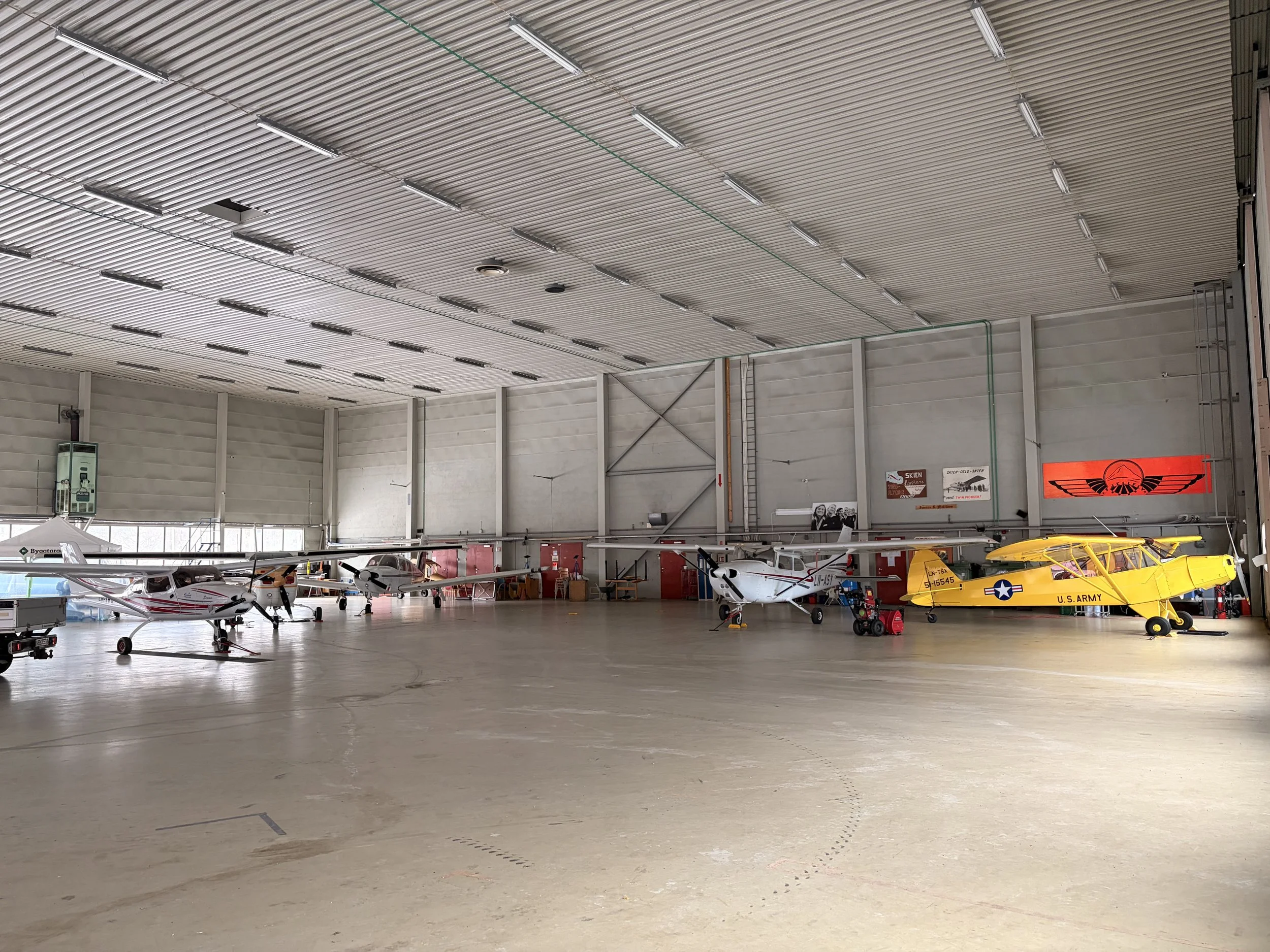 Indoor hangar with several small aircraft, including a yellow plane with U.S. Army markings, parked on a light-colored concrete floor.
