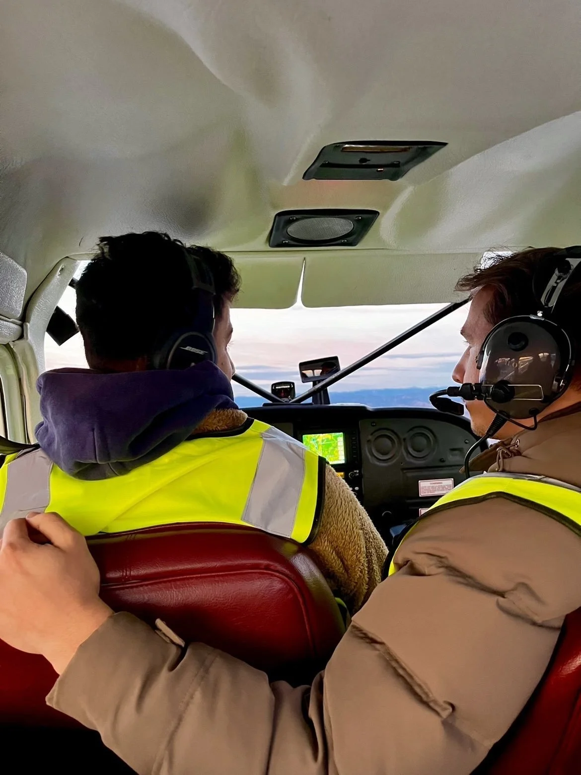 Two men wearing headphones and high-visibility vests sitting inside a small aircraft cockpit, with a view of the sky and clouds outside