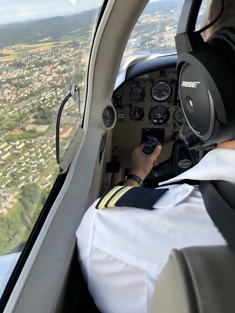 Utsikt fra cockpit av et lite fly som flyr over et grønnt landskap med boligområder.