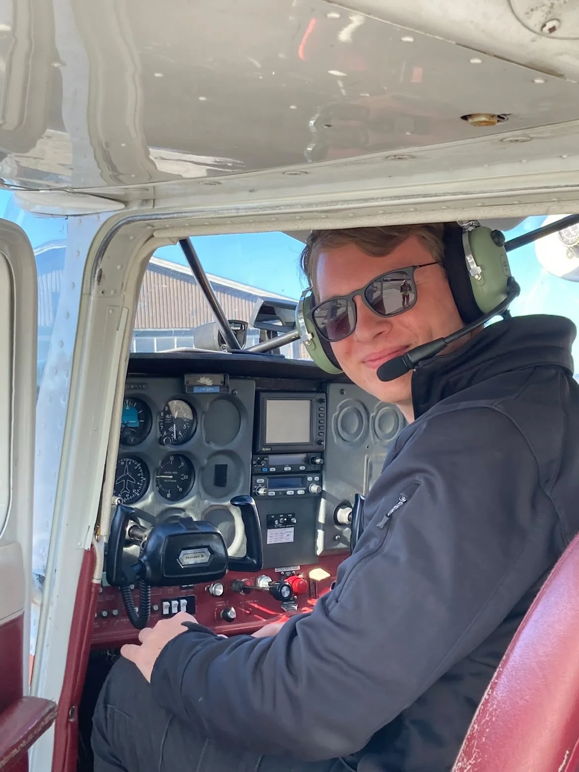 Young man sitting in the cockpit of a small airplane, wearing sunglasses and a headset, smiling at the camera.