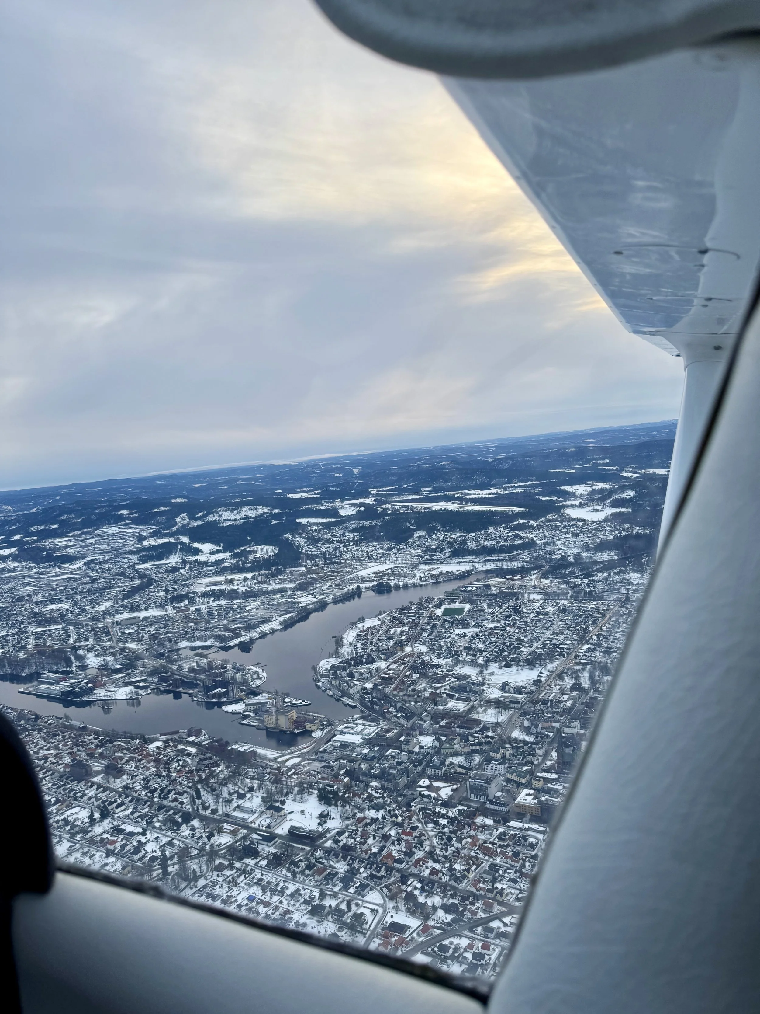 Flyfoto av en vinterby med elv, snødekte hus og veier sett fra et flyvindu