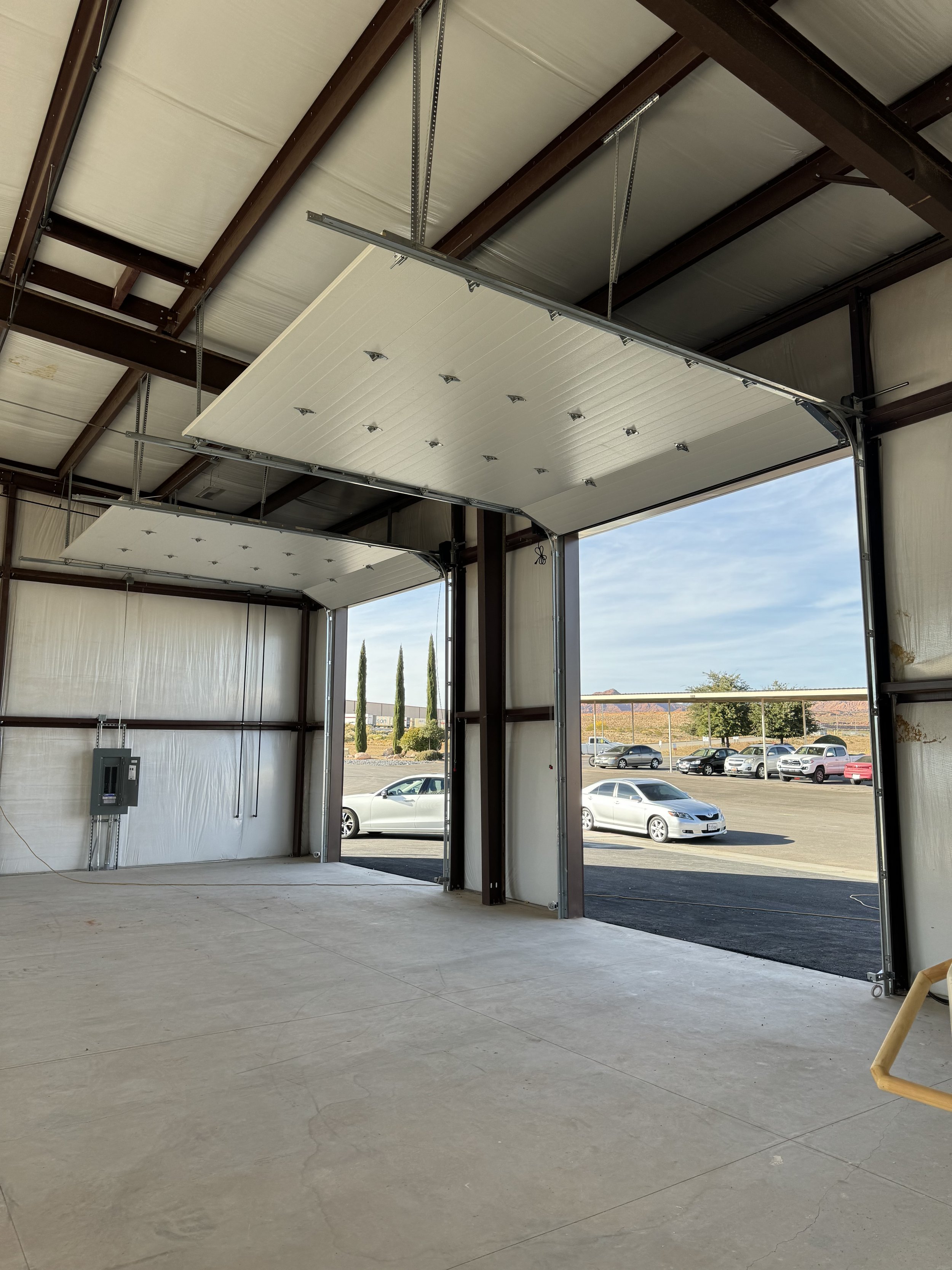 Inside a partially constructed commercial garage with two automatic garage doors open to an outside parking lot, a few trees, and a partly cloudy sky.