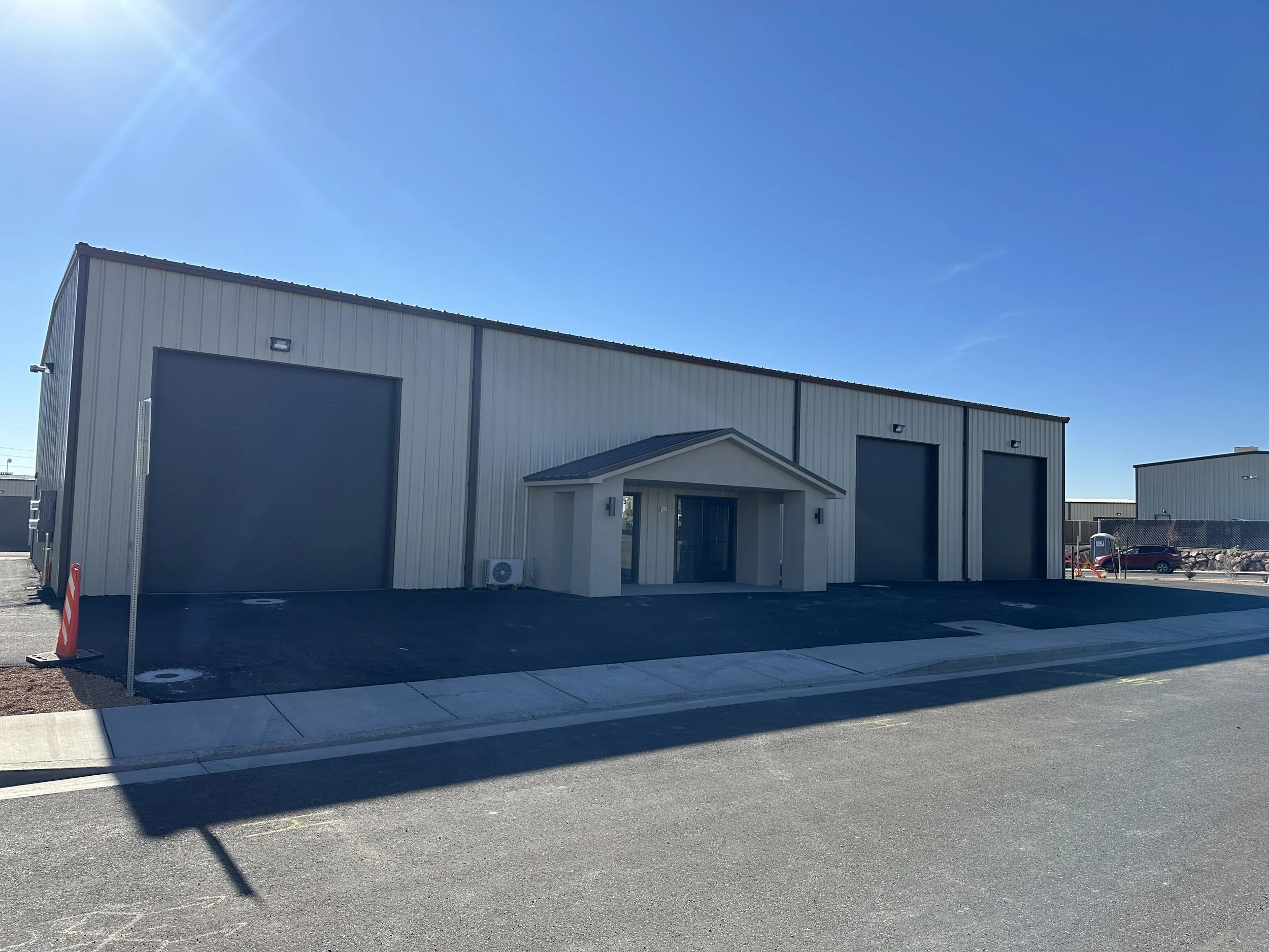 Commercial warehouse building with two large garage doors, a smaller entryway with a gabled roof, and an air conditioning unit outside. Clear blue sky and parked cars nearby. Hurricane UT