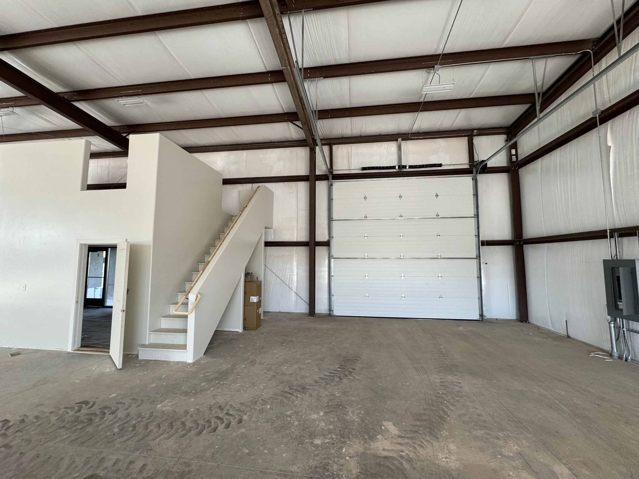 Interior of a large, unfinished warehouse with exposed metal beams and a high ceiling. There is a white partition with stairs leading to an upper level on the left side, and a large garage door at the back. The floor is dirt with tire marks. Some car