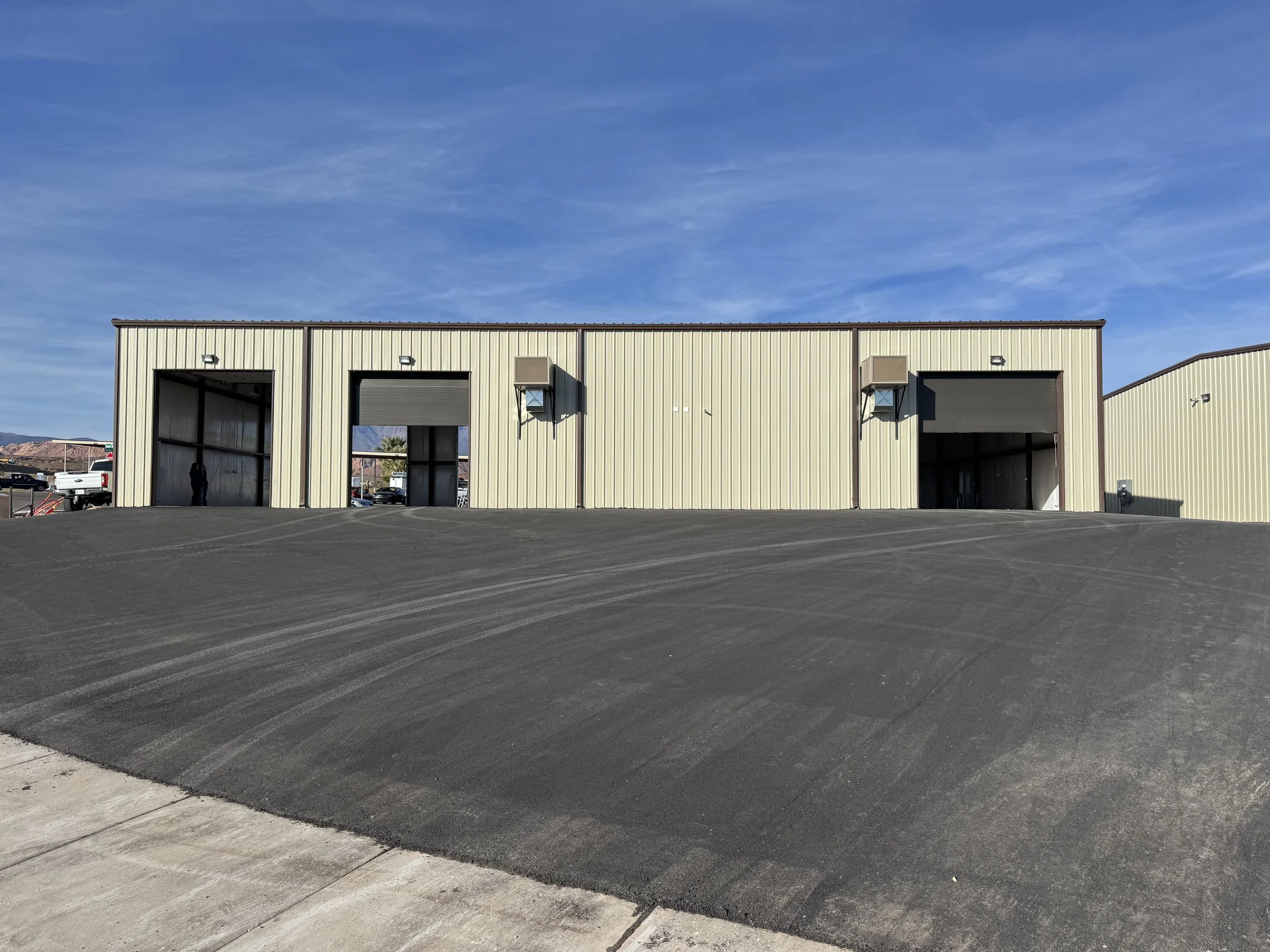 Large beige industrial warehouse building with three garage doors, two open and one closed, under a blue sky with some clouds. The foreground shows freshly paved black asphalt.
