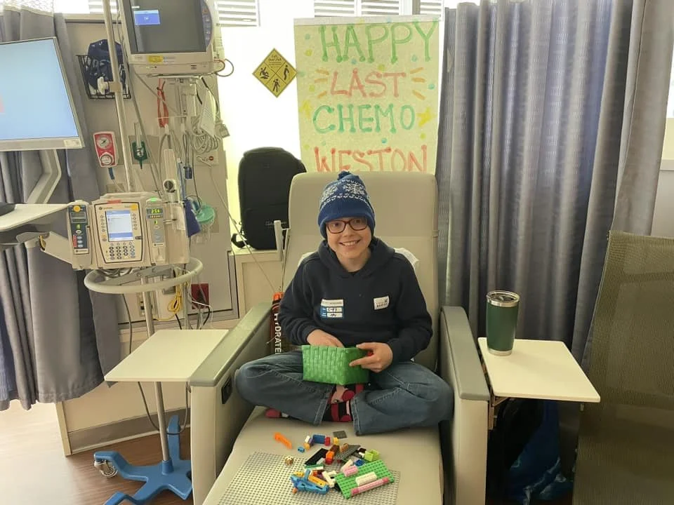 A smiling young girl sitting cross-legged in a hospital bed, wearing a blue knit hat, glasses, and a dark hoodie, holding a green Lego piece. There are Lego bricks on the bed and a sign in the background that says 'HAPPY LAST CHEMO KIeston.' Medical 