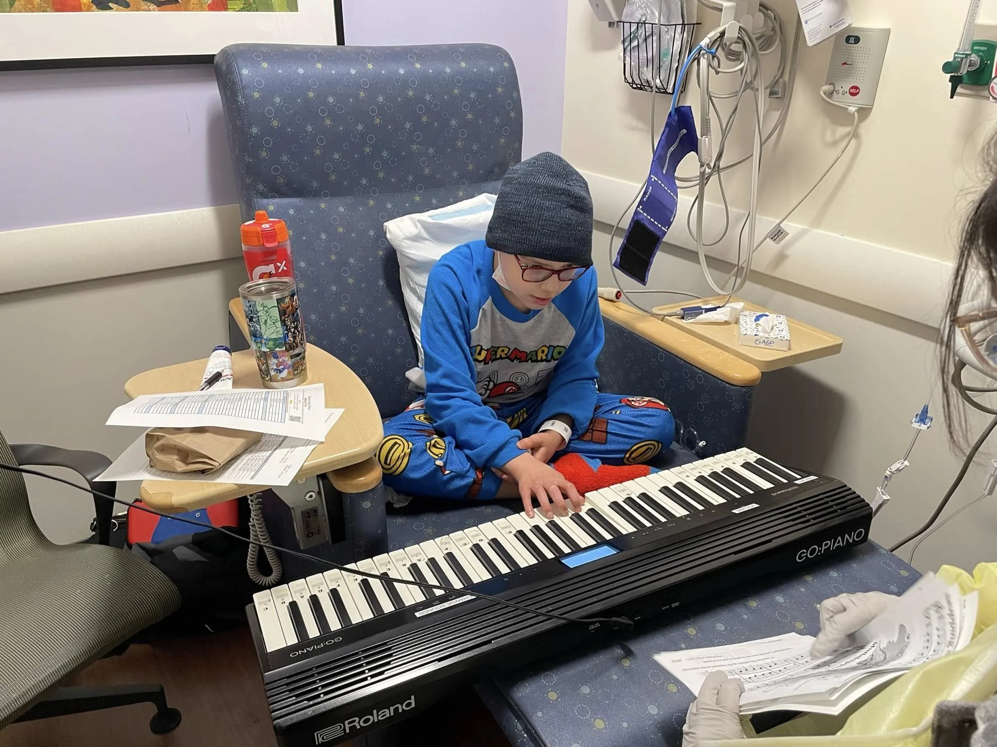 A young boy sitting cross-legged on a hospital bed playing a keyboard piano, wearing glasses, a beanie, and blue pajamas with video game characters. There are medical monitors and cords on the wall nearby, and a doctor holding a sheet of paper and we