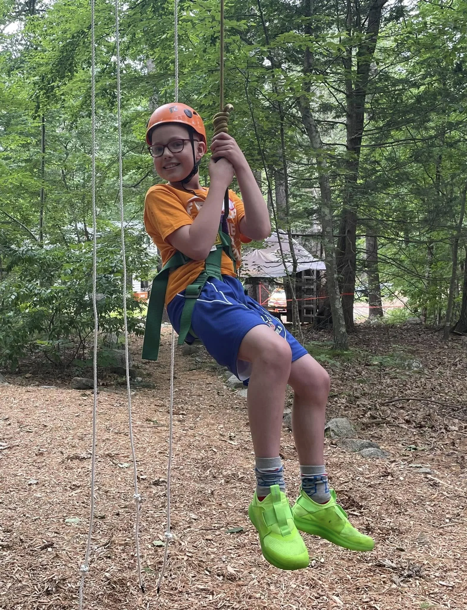 A young boy wearing glasses, a safety helmet, and bright green athletic shoes, sitting on a swing in a wooded area, holding onto the ropes with a harness and smiling at the camera.