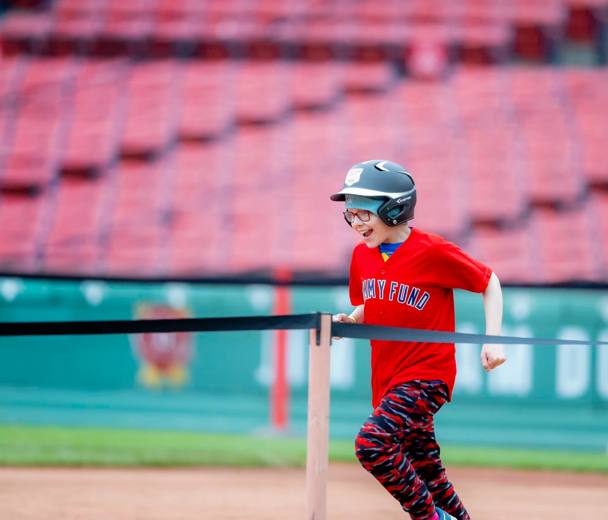 Young child wearing a batting helmet and glasses, running on a baseball field with empty stands in the background.