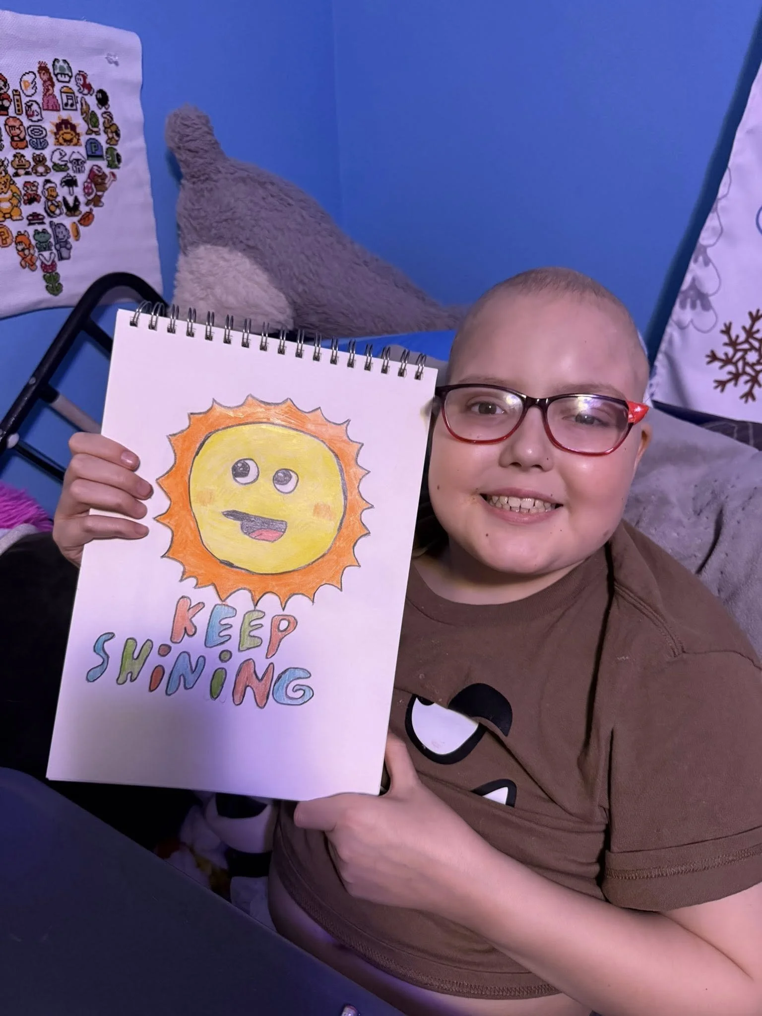 A smiling person with glasses holding a drawing of a smiling sun with the text 'Keep Shining' beneath it.