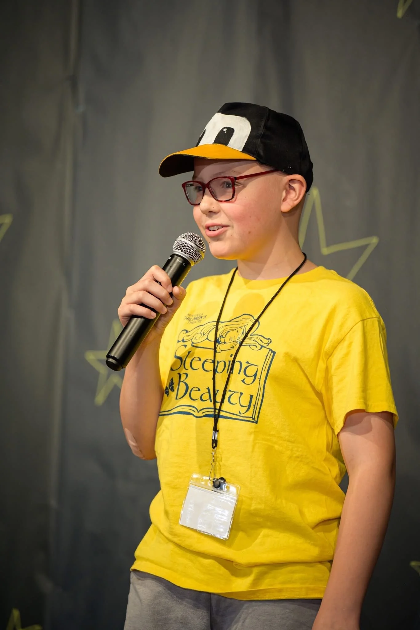 A young person wearing a yellow t-shirt with the words 'Sleeping Beauty', red glasses, and a black and yellow penguin-themed cap, speaking into a microphone.