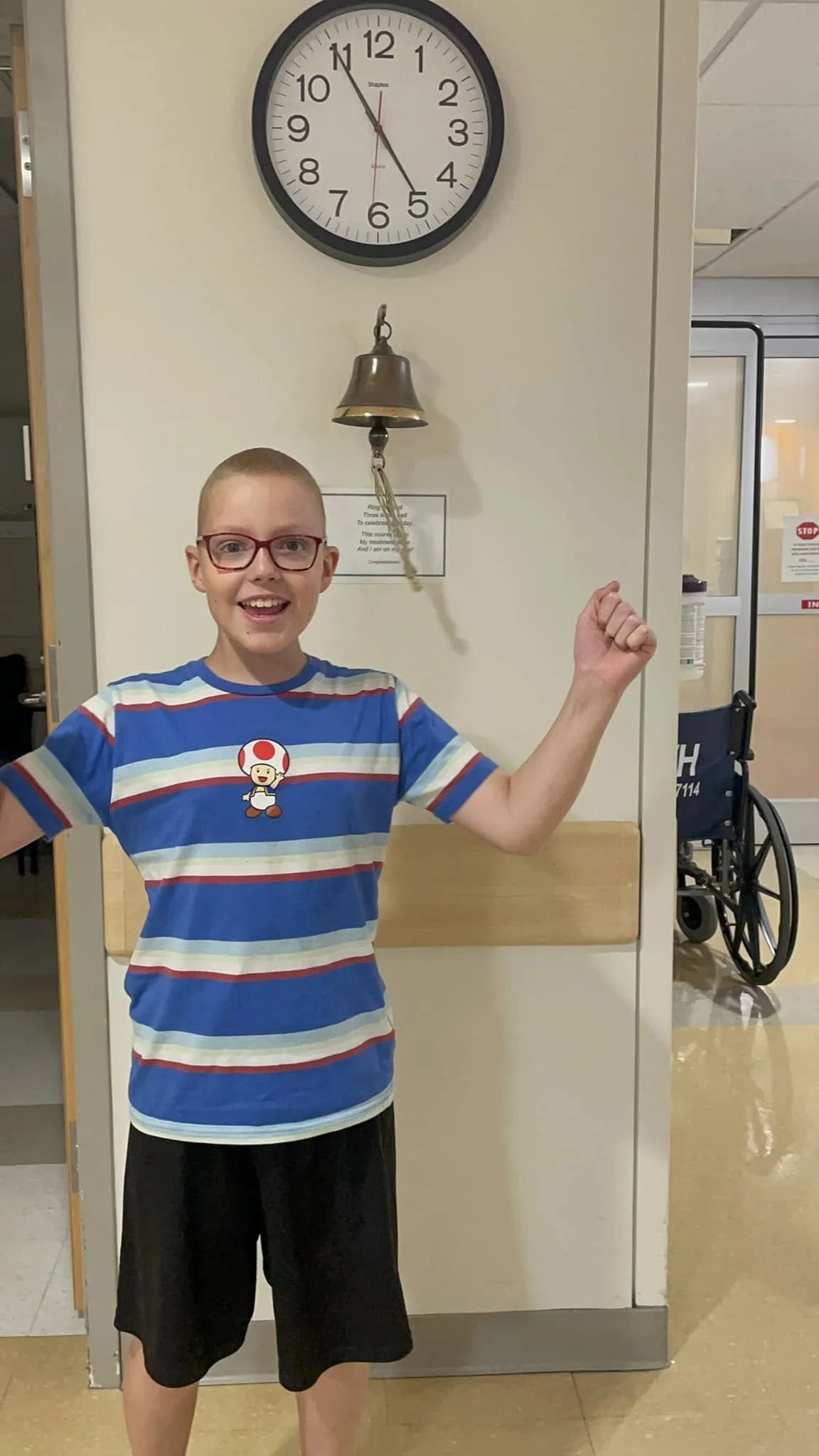 A smiling boy wearing glasses and a blue striped T-shirt standing in front of a hospital wall with a bell hanging on it. A clock shows the time as 10:55. There is a wheelchair in the background.