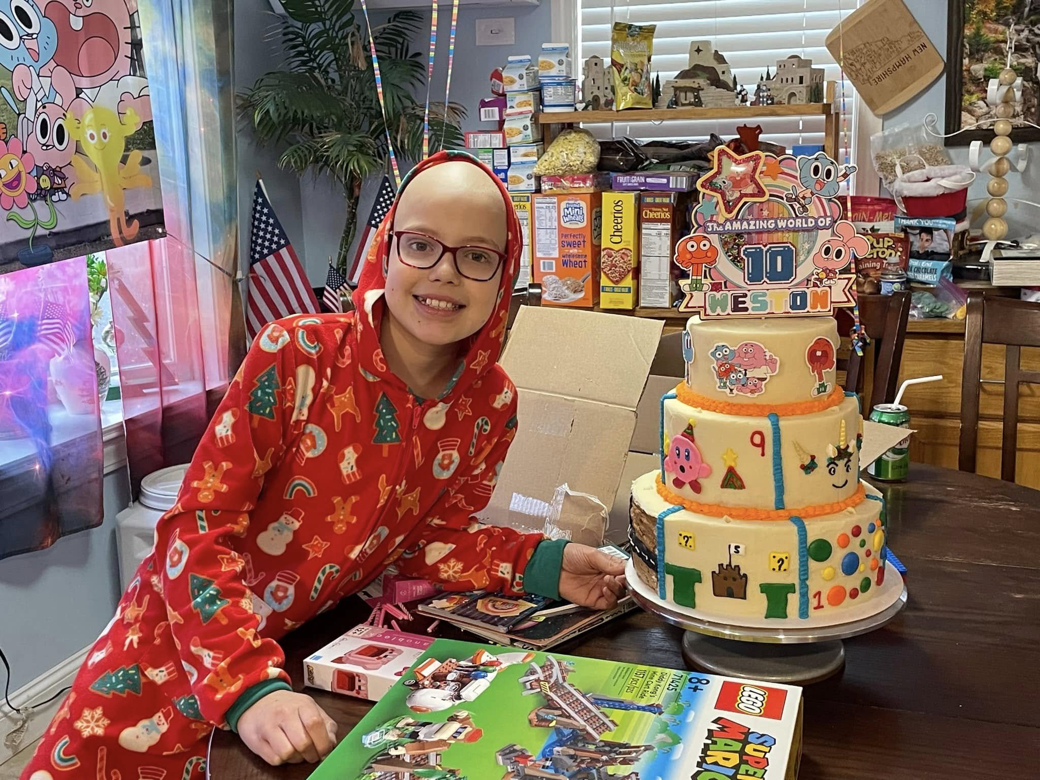 A girl with glasses smiling at a birthday party, dressed in a red Christmas-themed hoodie, sitting next to a decorated cake featuring Lego and Super Mario themes, on a wooden table surrounded by several presents and party decorations.