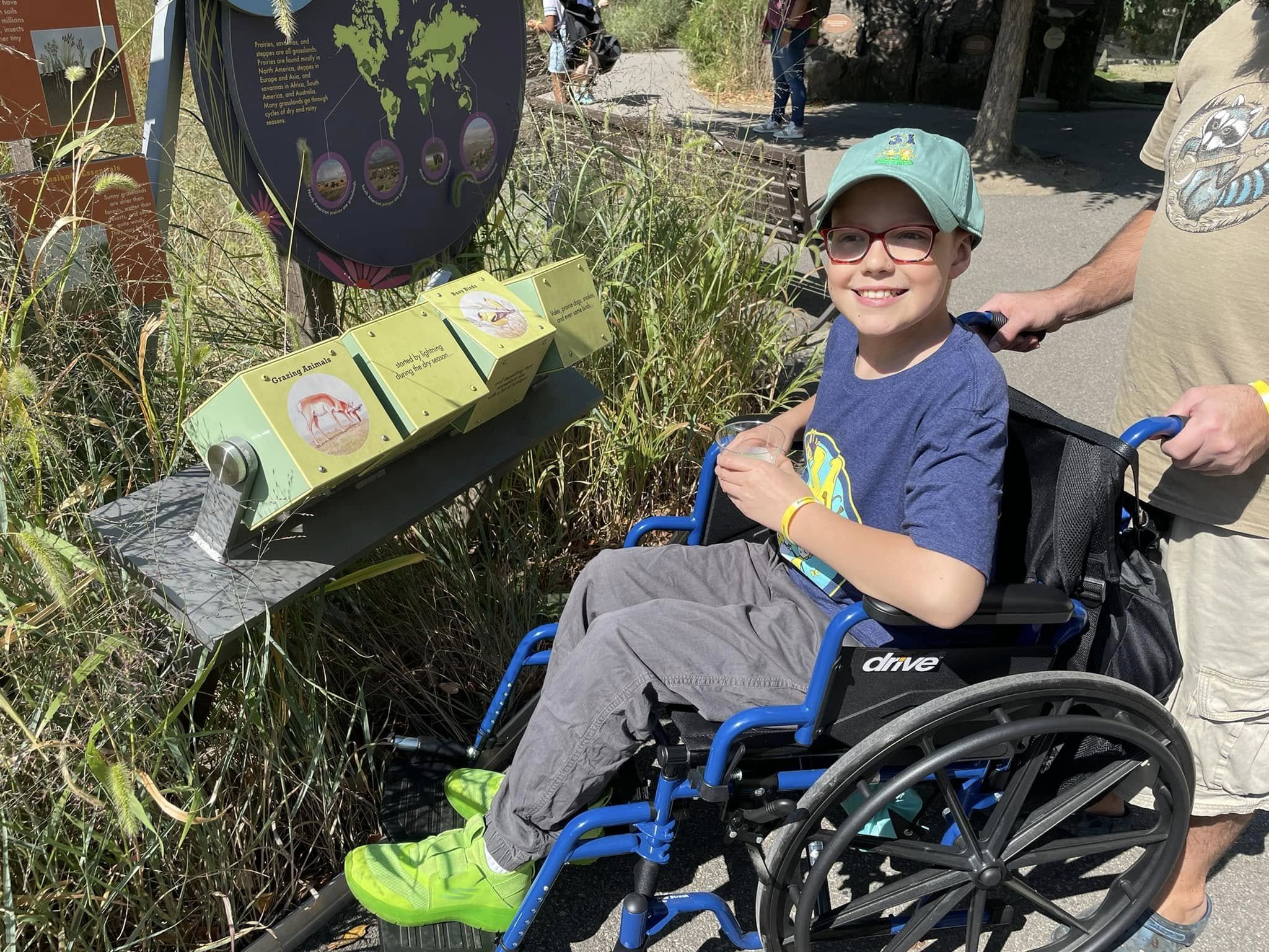 A young boy with glasses and a green hat sitting in a wheelchair, smiling at an educational display about animals at a park or zoo, with an adult man standing behind him.