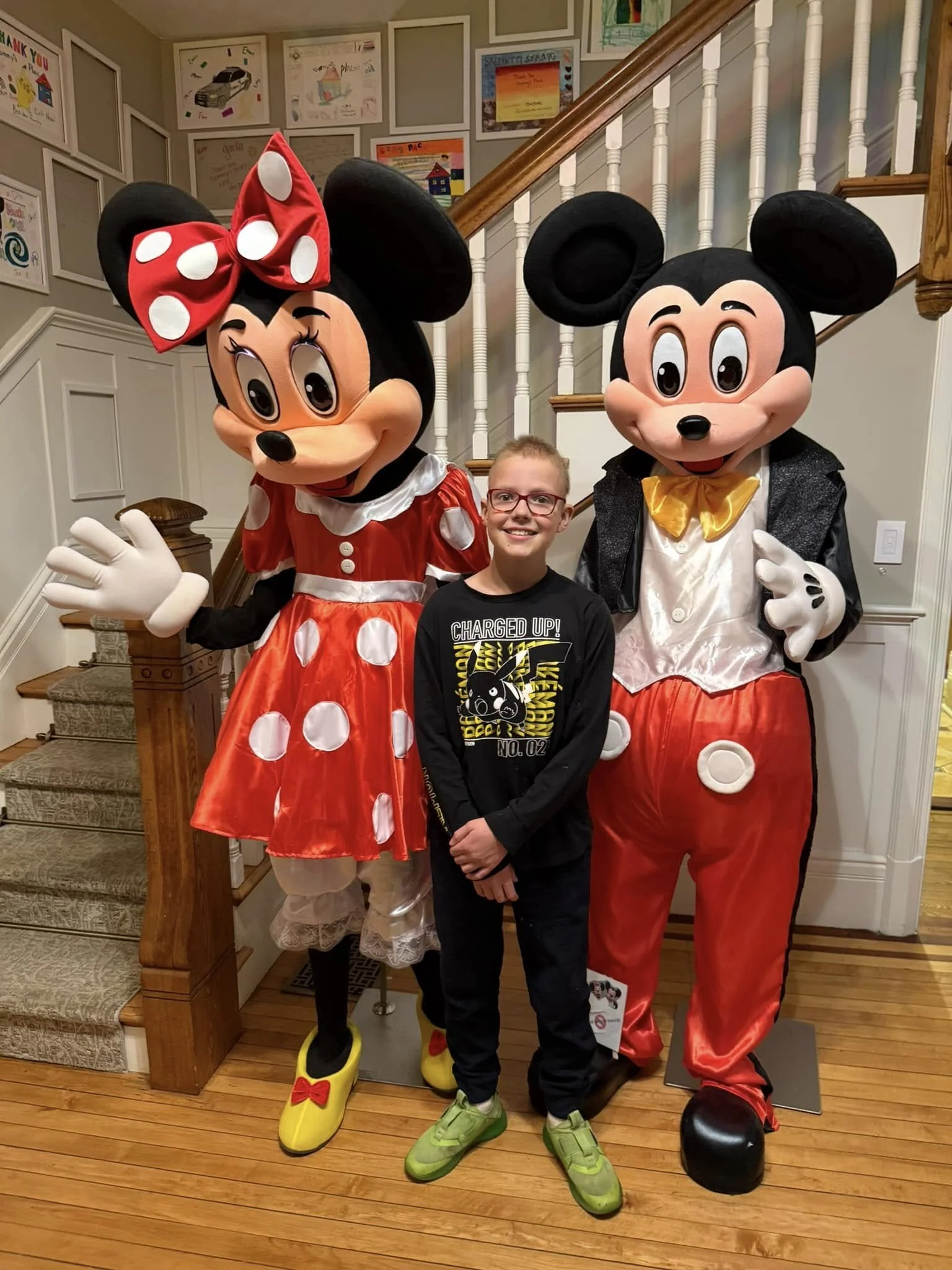 A child with glasses and a black shirt stands between life-sized Mickey and Minnie Mouse costumes inside a house with framed artwork on the walls and a wooden staircase in the background.