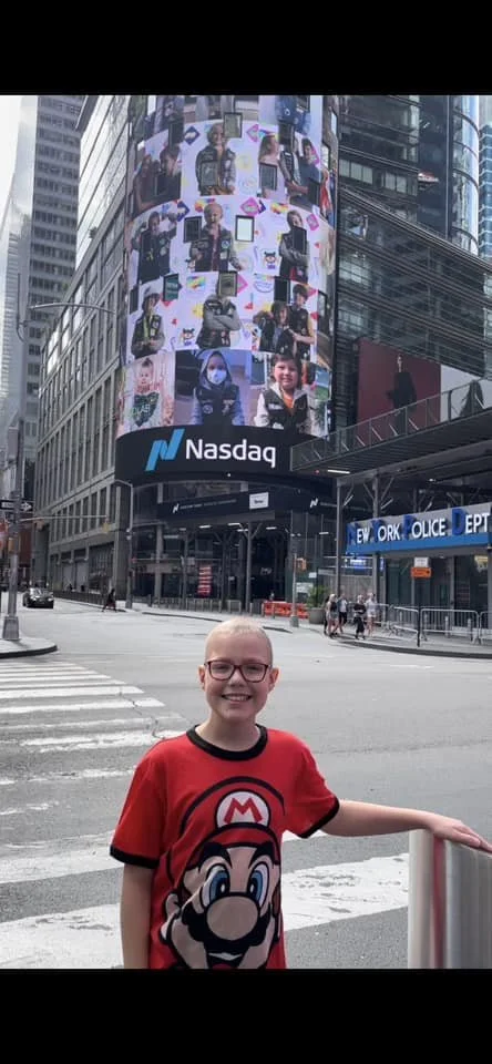 A young boy wearing a red Mario t-shirt smiling at the camera, standing in Times Square with a large digital Nasdaq billboard displaying various images of people in the background.