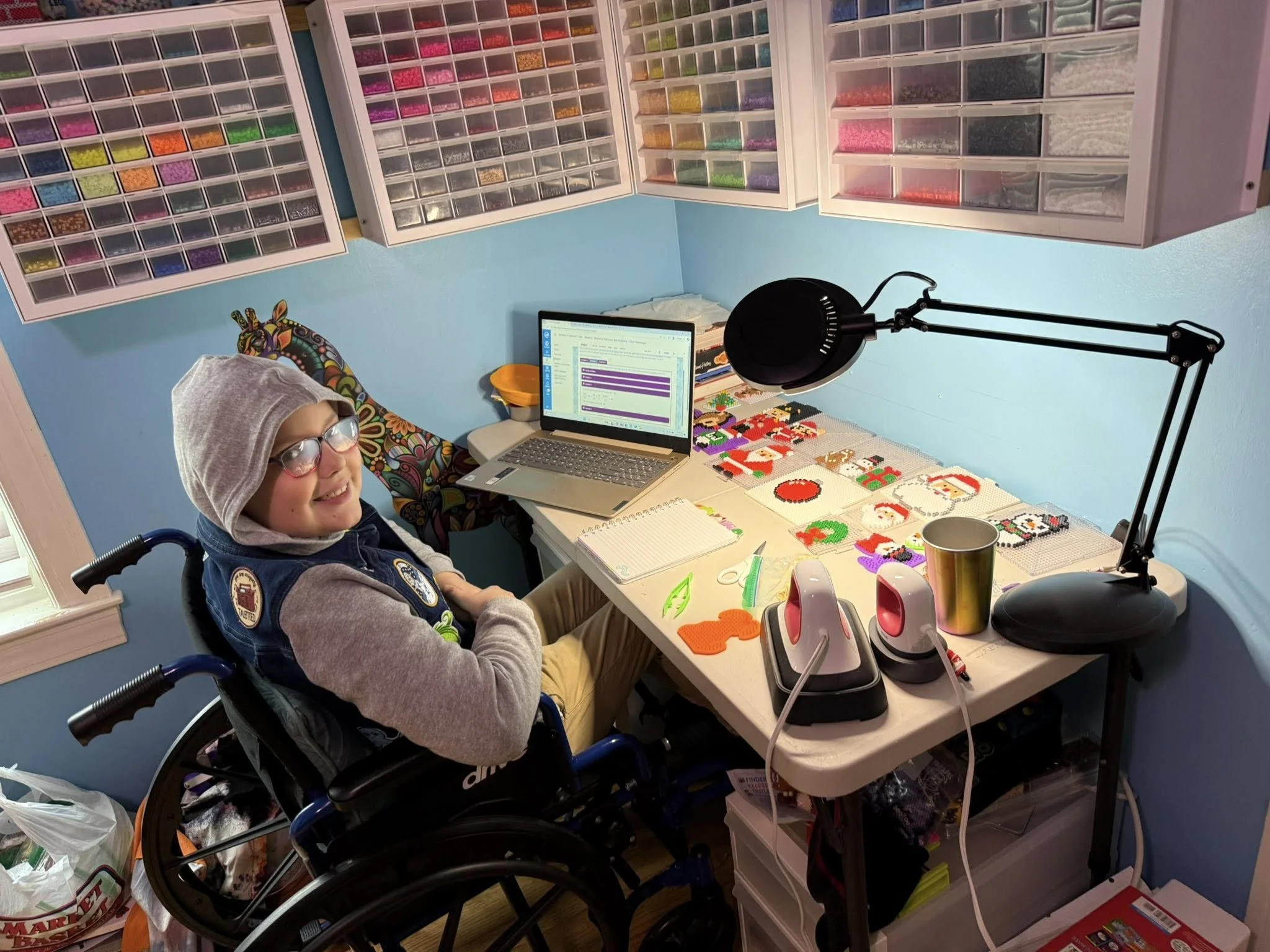 A young person in a wheelchair in a craft room, smiling at the camera. The workspace includes a computer, a lamp, a notebook, a mug, and various craft supplies like cross-stitched artwork of Christmas and holiday themes on the table, with organized s