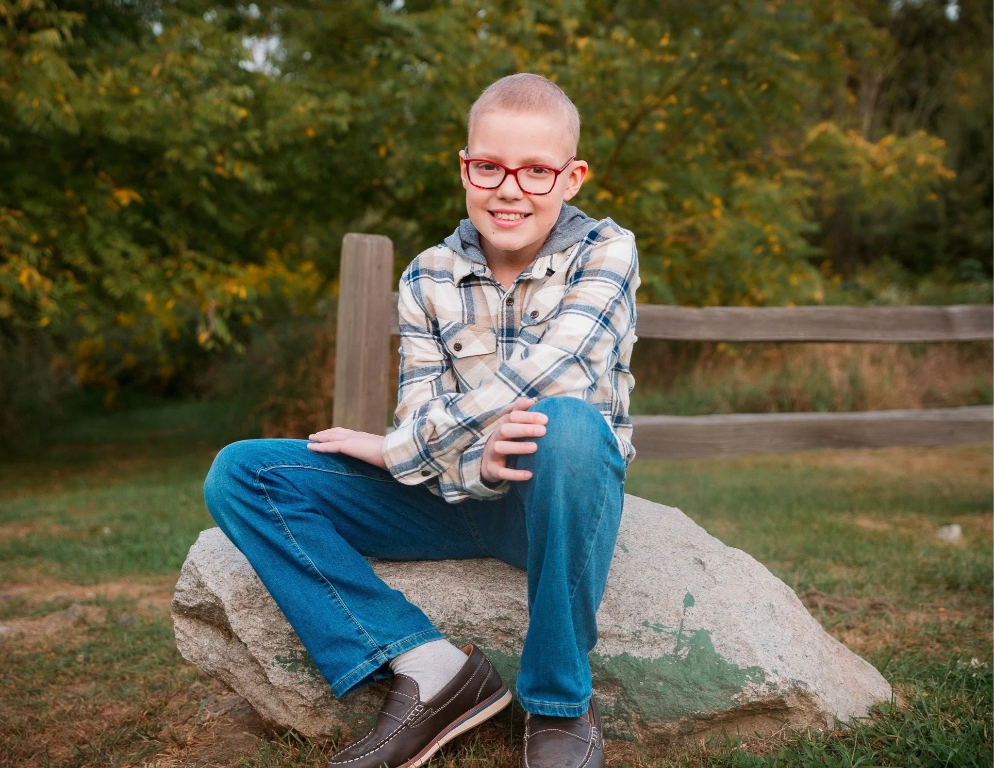 A boy with glasses and a plaid shirt sitting on a large rock outdoors during fall.